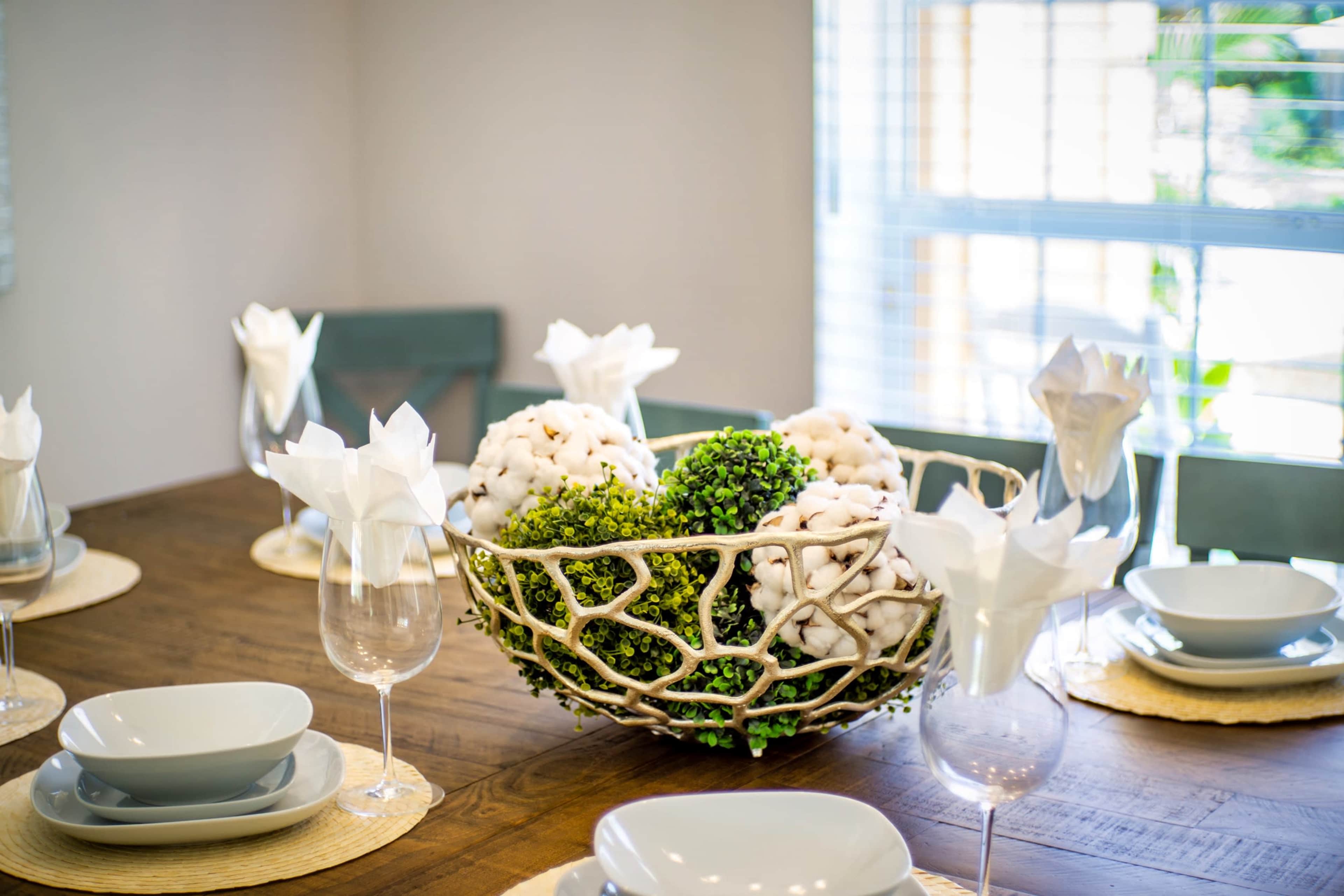 A dining table is set with grey plates and glasses, featuring a decorative bowl of cotton and greenery at the center.