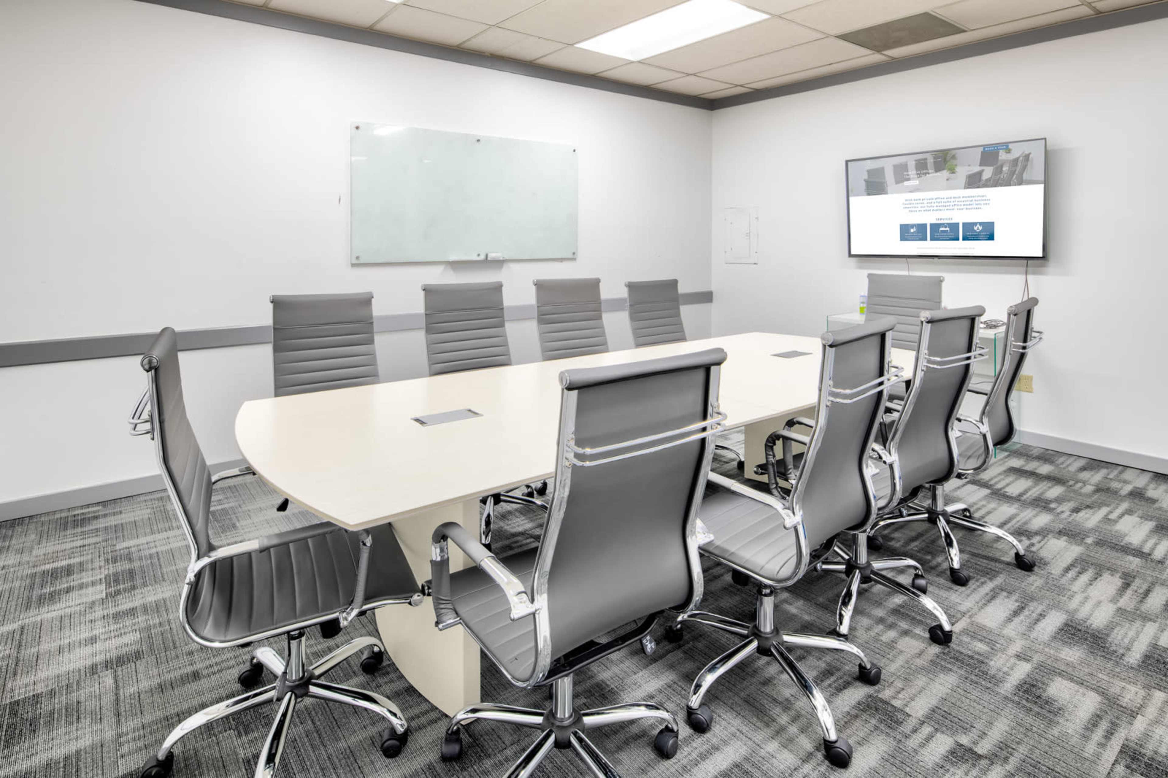 A modern conference room features a long table surrounded by ergonomic chairs, with a wall-mounted screen and a whiteboard.