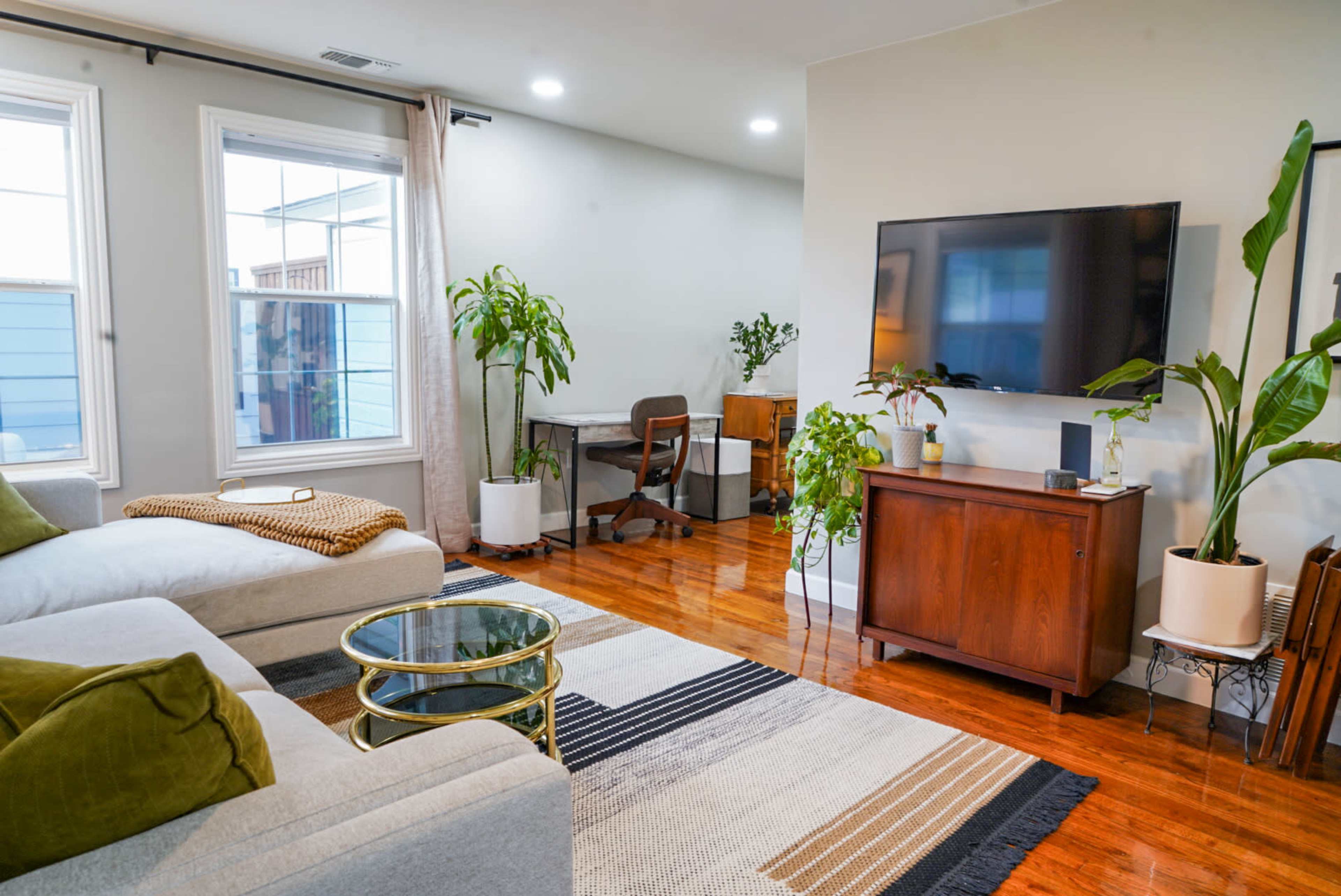 A modern living room with a sectional sofa, a glass coffee table, a desk area, and indoor plants, all positioned around wooden flooring and natural light from windows.