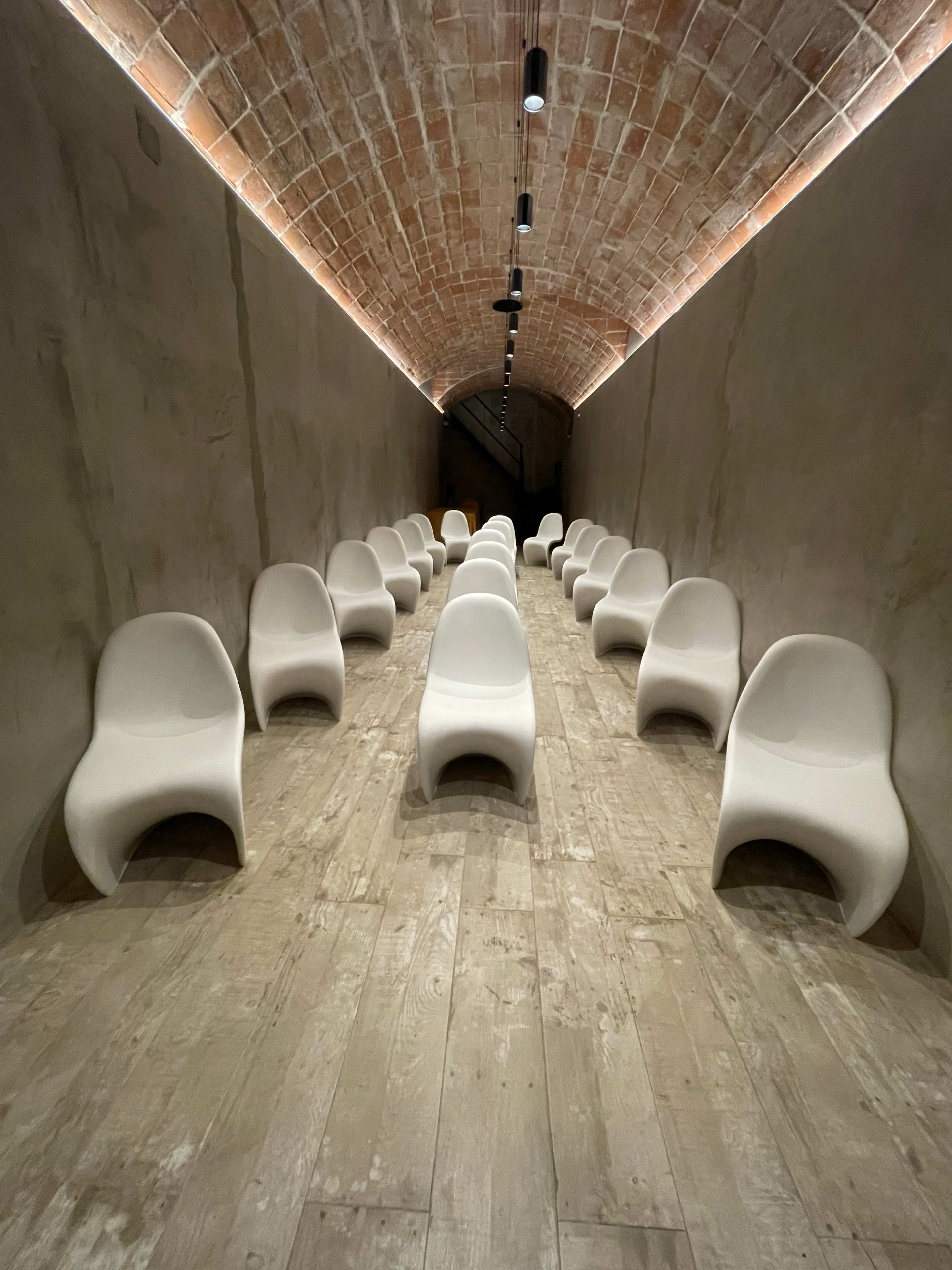 A long, narrow hallway featuring several white chairs arranged in rows on a wooden floor, with a brick archway ceiling above.