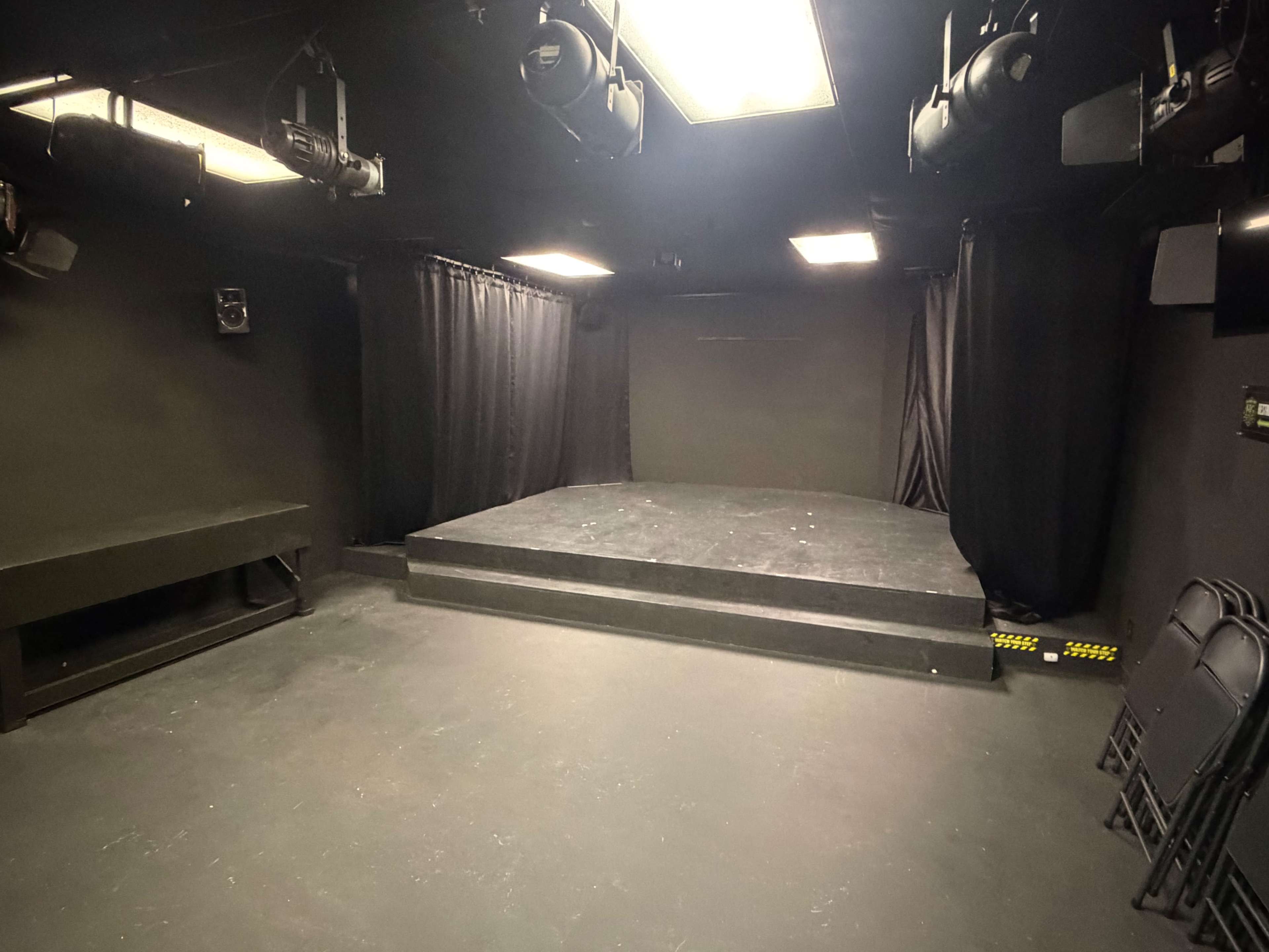The image shows a small, dimly lit theater space with a raised stage, black curtains, and several folding chairs stacked against the wall.