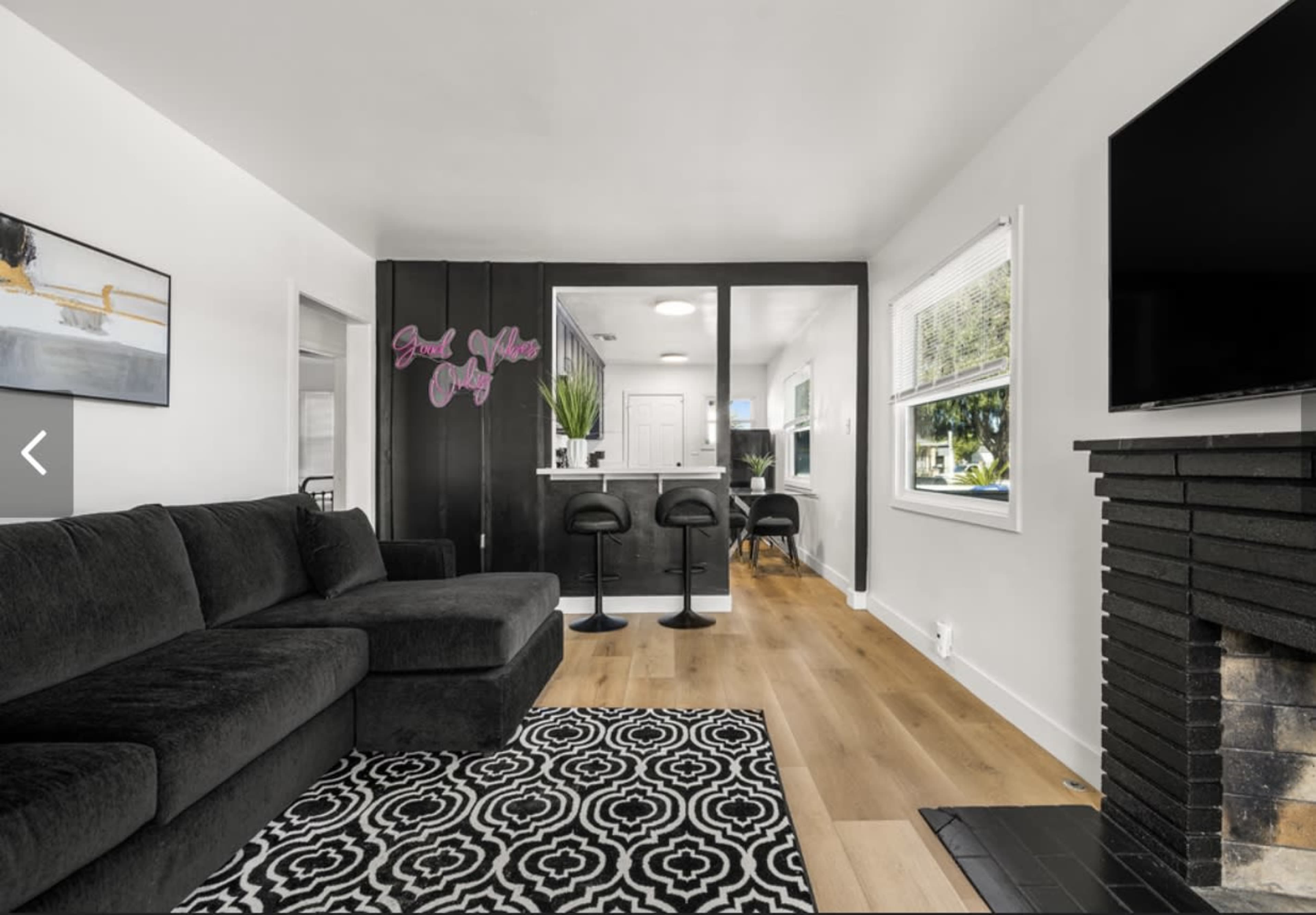 A modern living room features a black sectional sofa, a patterned rug, and a view into a kitchen area with bar stools and a window.