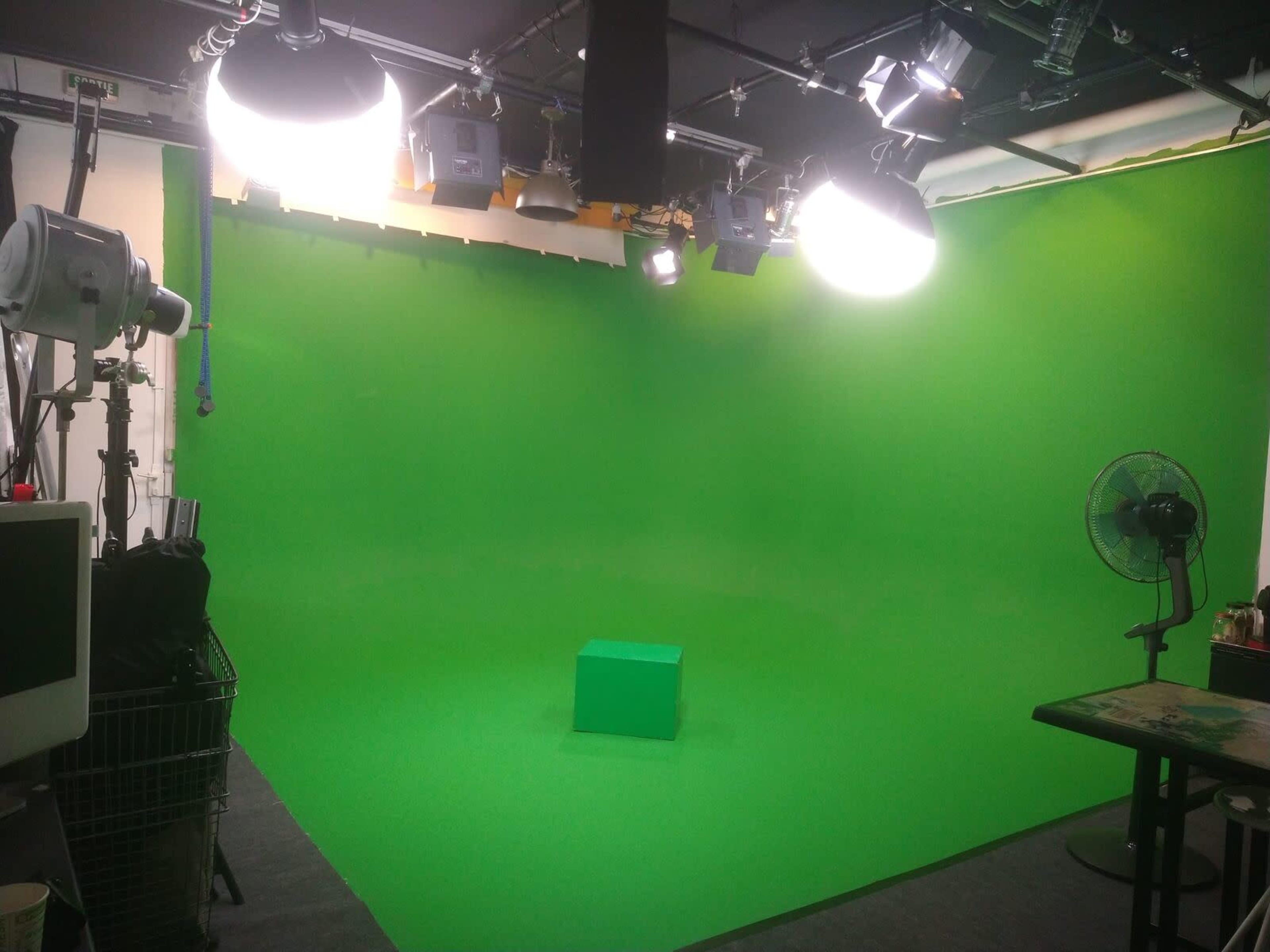 The image shows a green screen studio with a single green block in the center, surrounded by overhead lights and a fan.