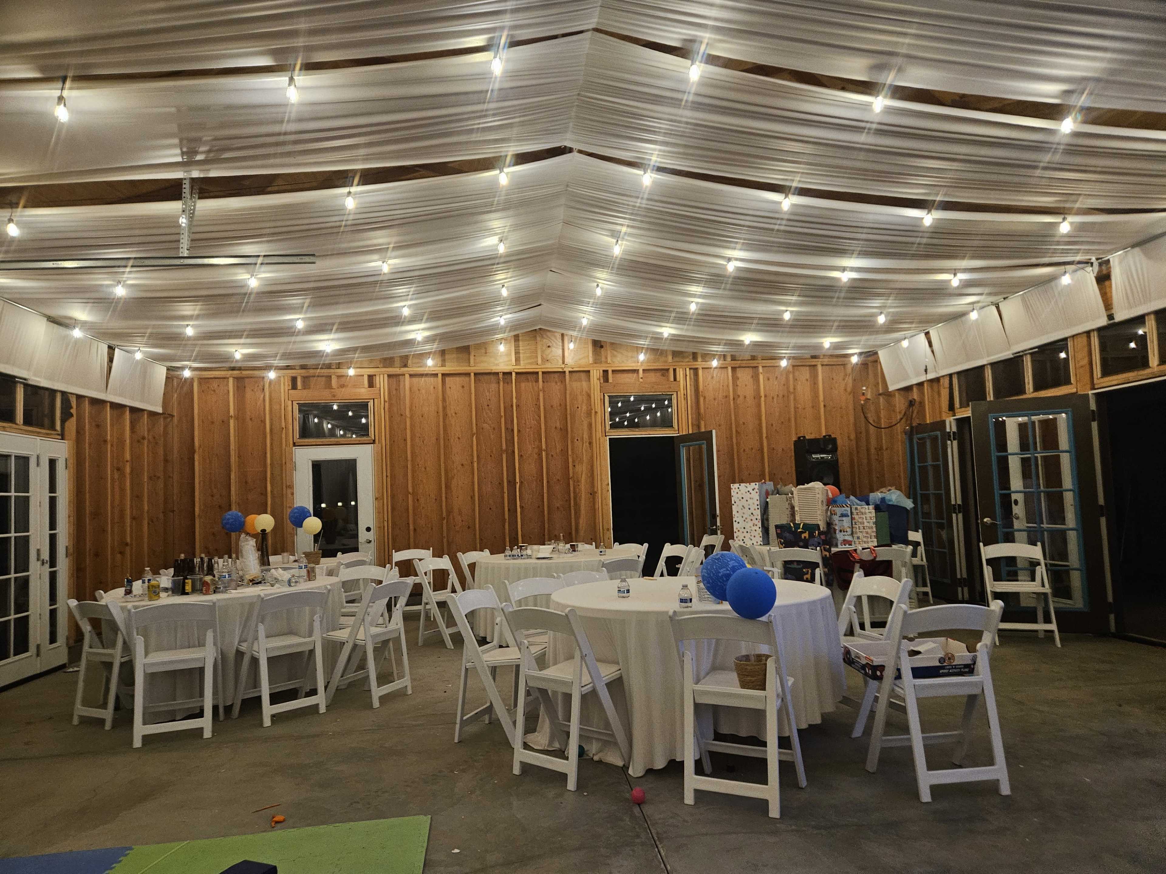 The image shows a large, well-lit event space with several round tables covered in white tablecloths, surrounded by white chairs, and decorated with balloons and gift packages.