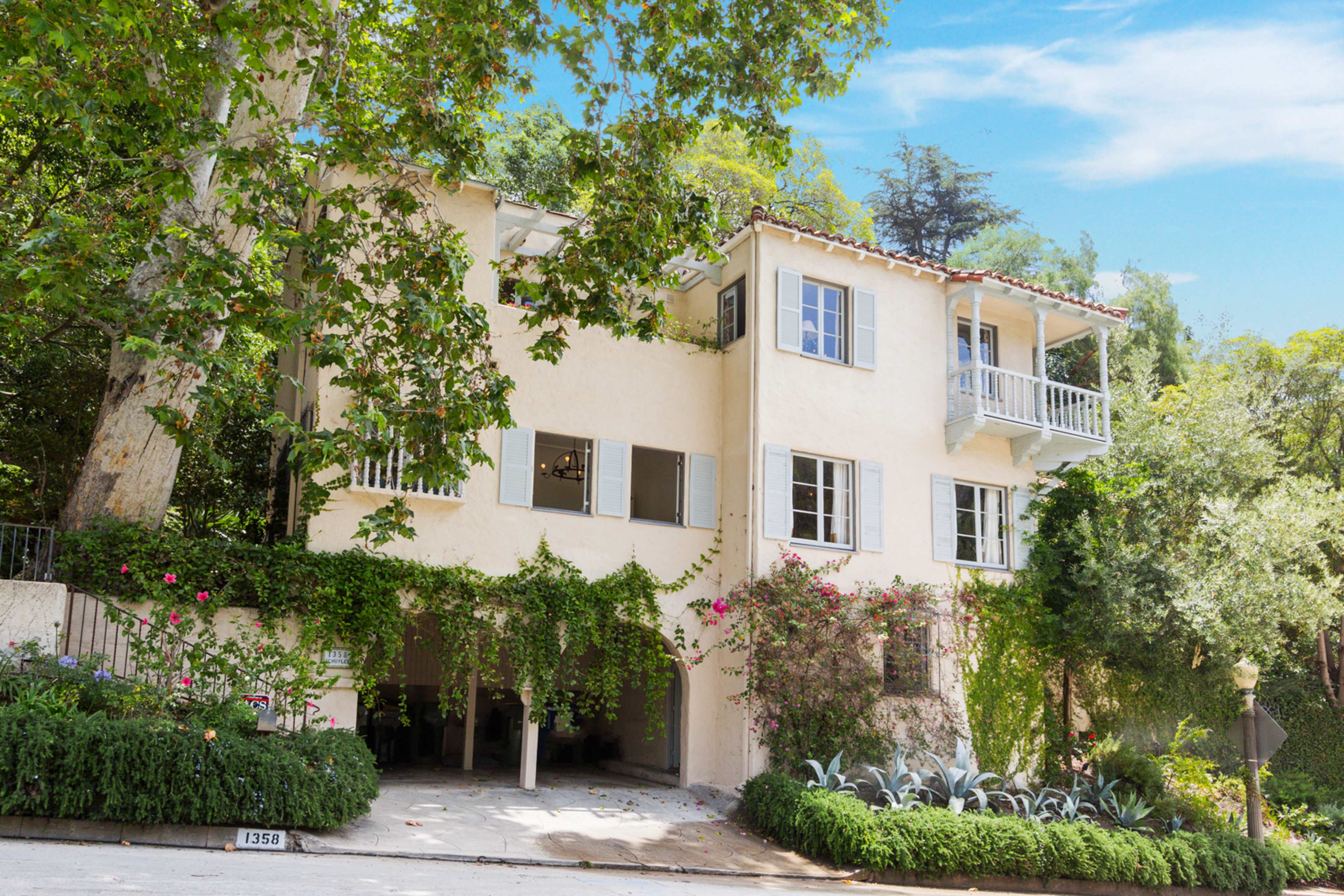 A three-story house with a beige exterior, surrounded by greenery and flowers, features multiple windows and balconies.