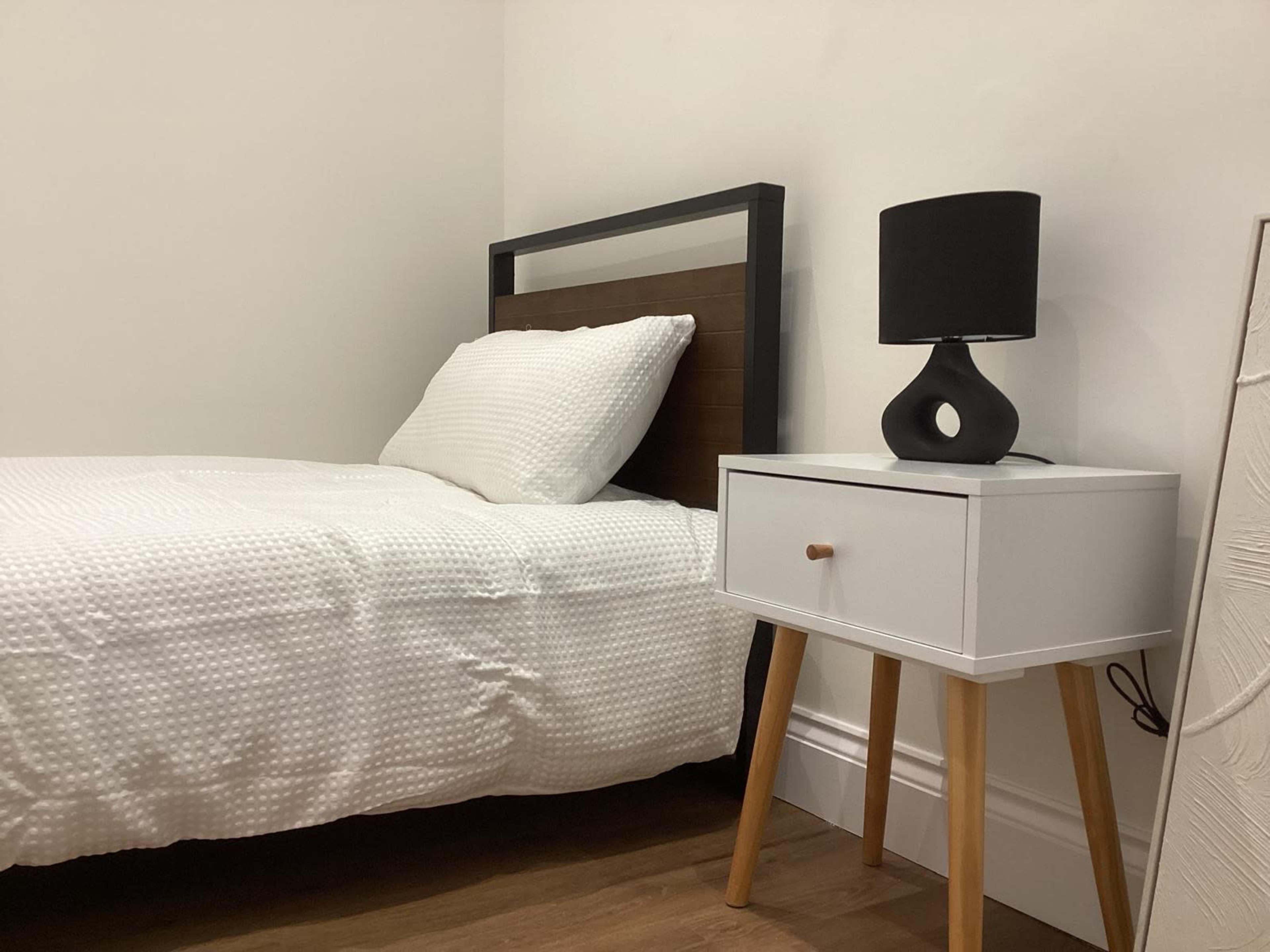 The image shows a simple bedroom featuring a bed with a white dotted blanket, a bedside table with a black lamp, and wooden flooring.