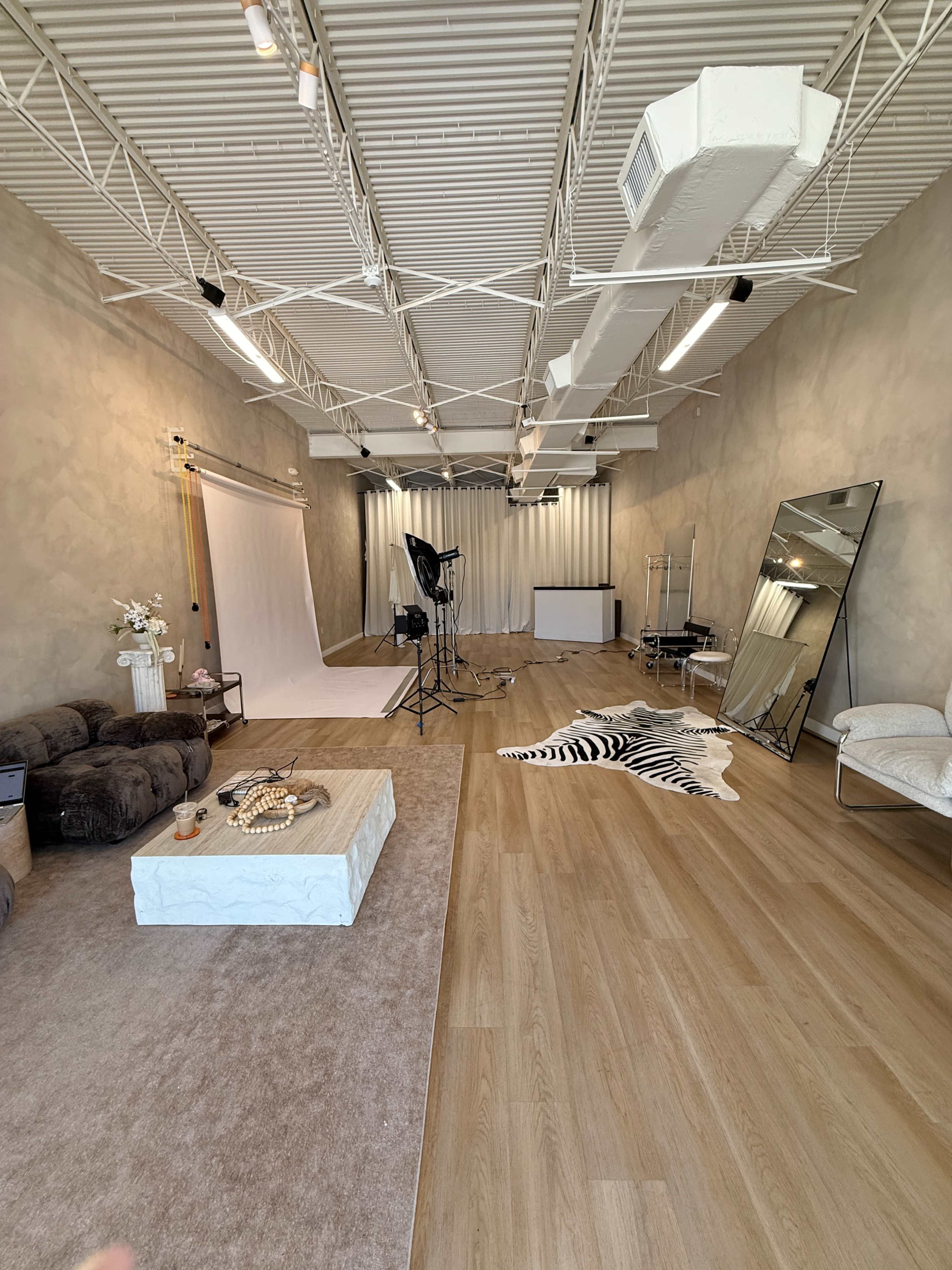 The image shows a spacious photography studio featuring a backdrop setup, lighting equipment, various seating areas, and a large mirror on a wooden floor.
