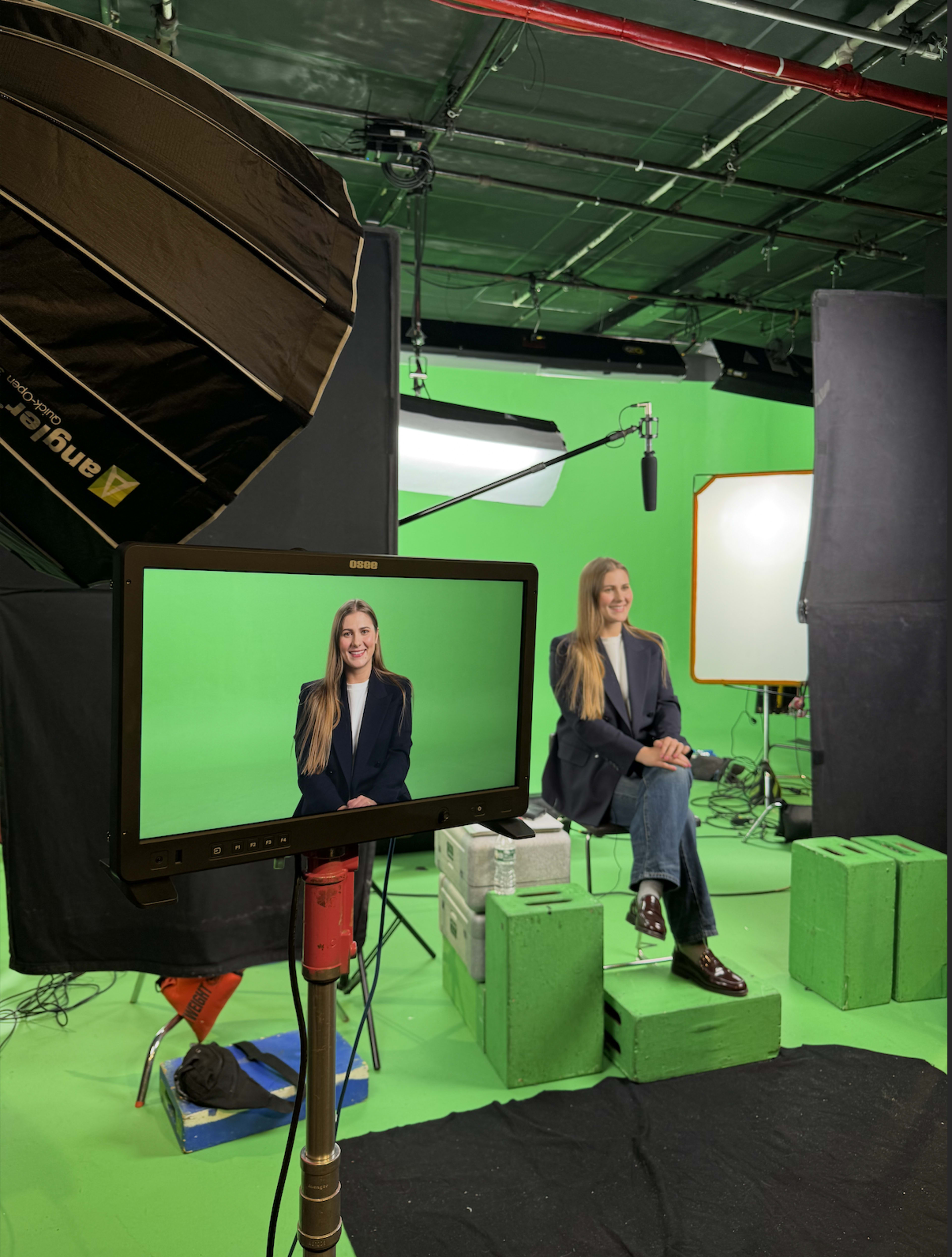 A person with long hair sits on a stool in front of a green screen, while being filmed and lit by studio equipment.