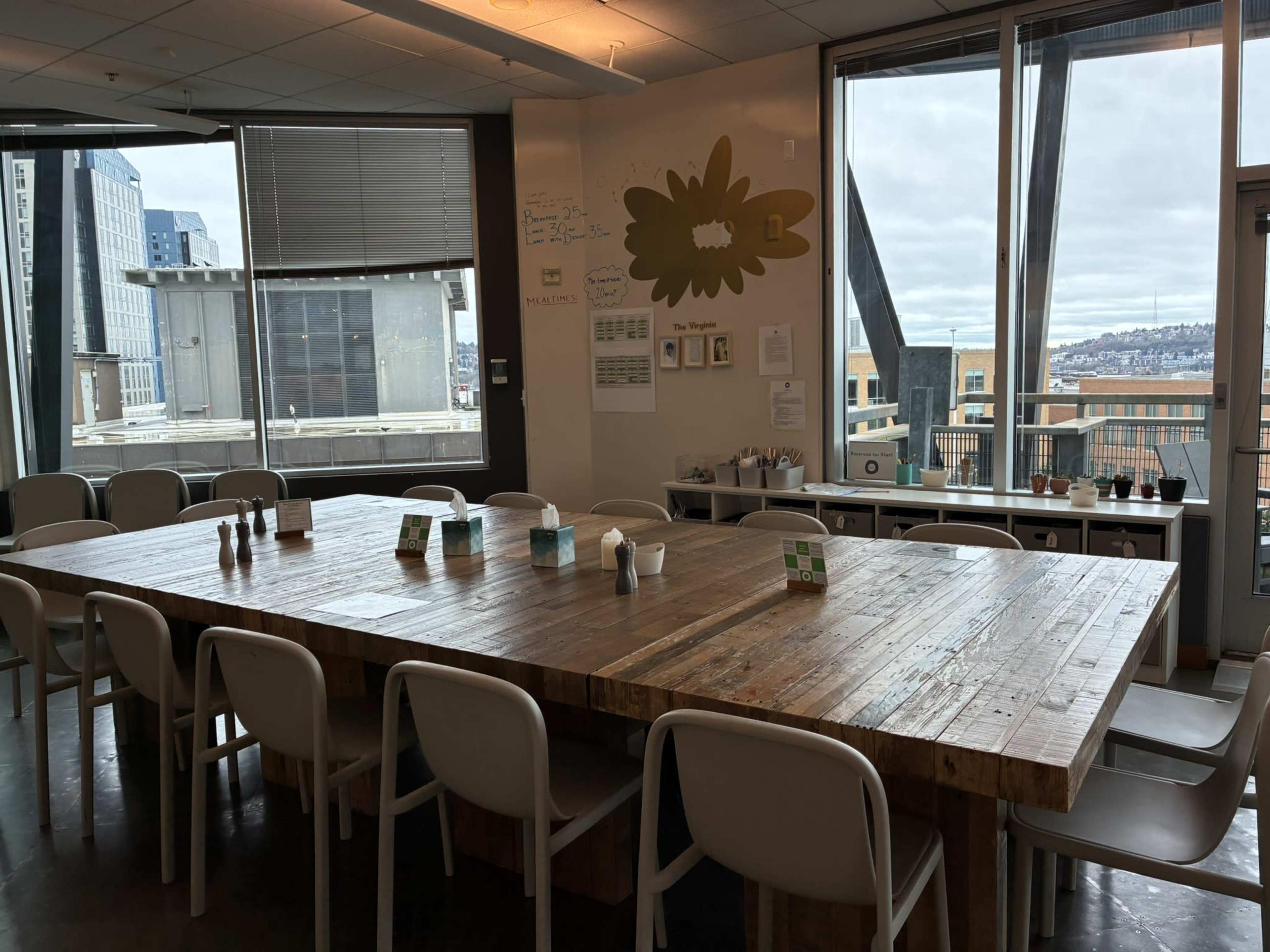 A large, wooden table surrounded by white chairs is set in a brightly lit room with windows overlooking an urban landscape.