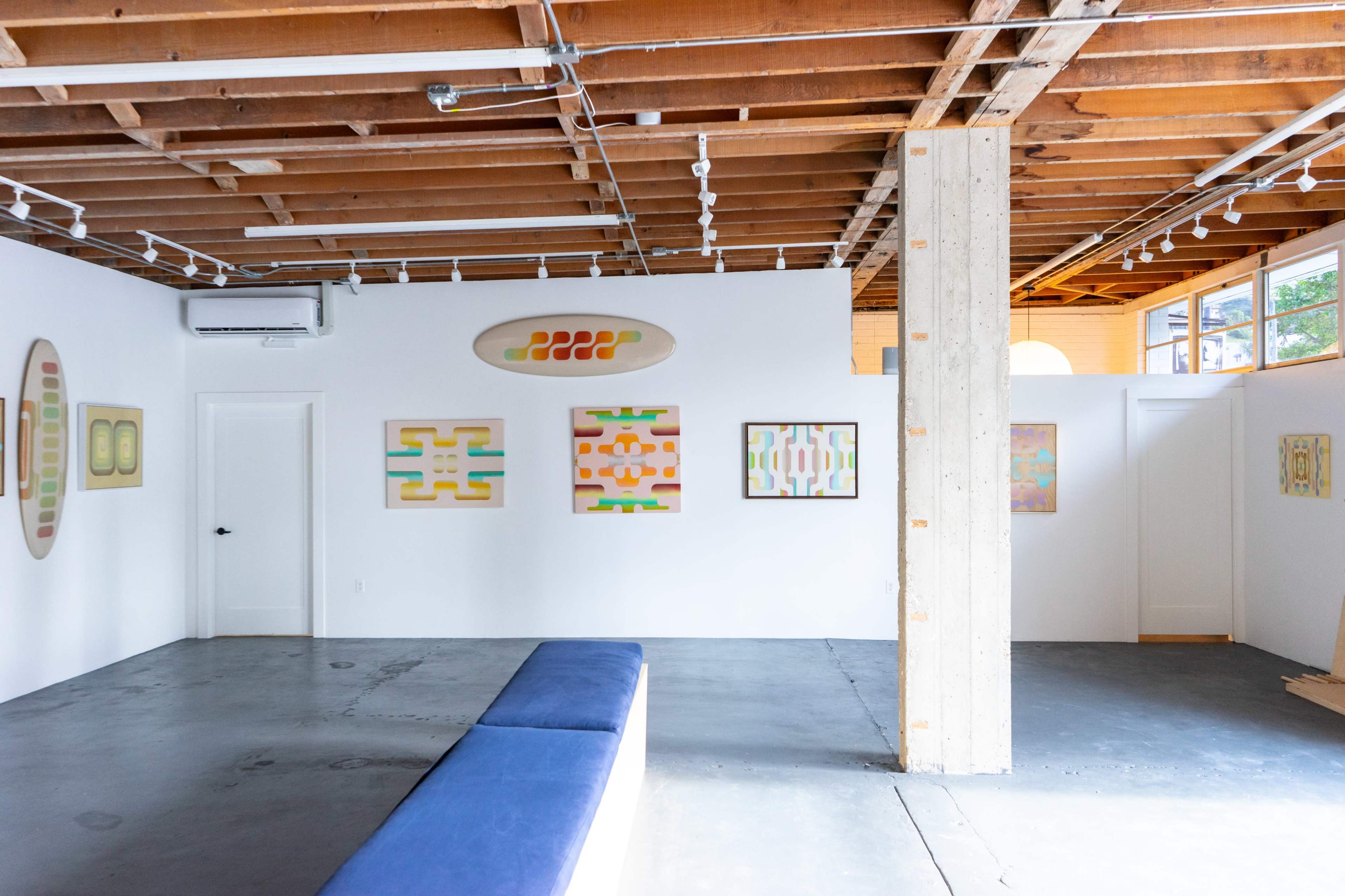 The image shows a spacious art gallery with white walls displaying several abstract artworks, a blue bench in the center, and wooden beams overhead.