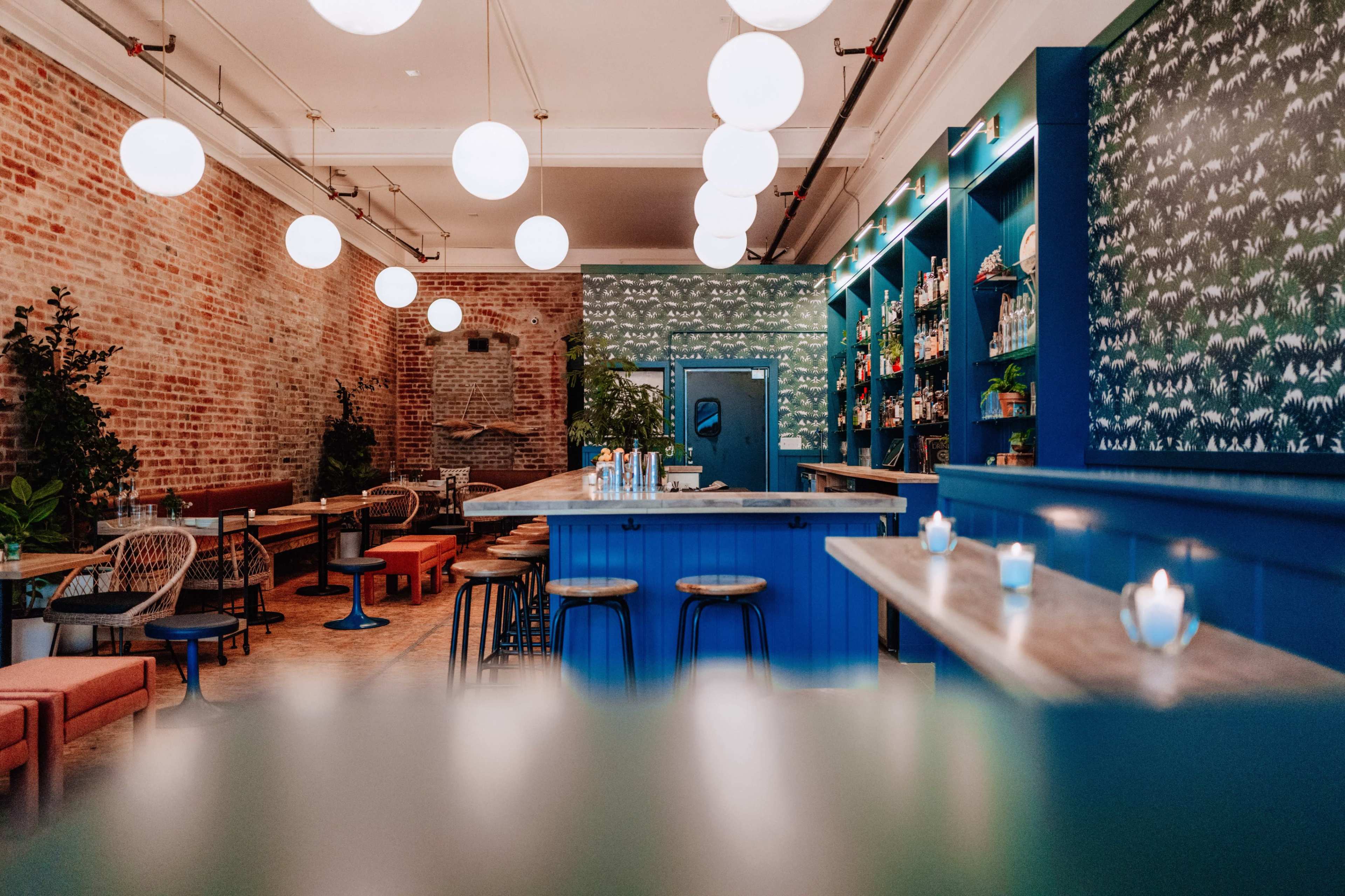 A modern bar interior with a blue counter and shelves, illuminated by round light fixtures, against a backdrop of exposed brick and patterned wallpaper.