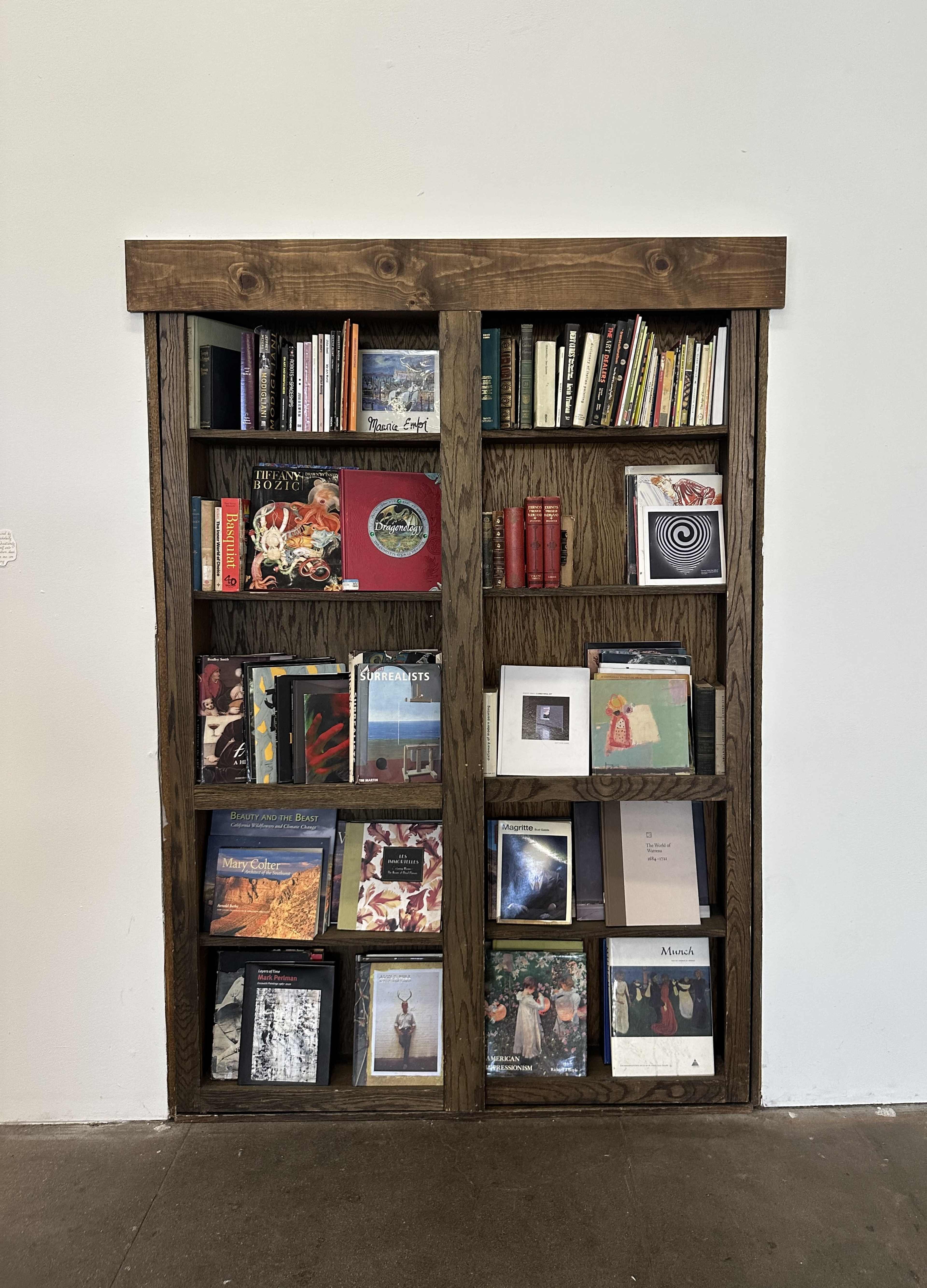 The image shows a wooden bookshelf filled with various books and albums organized in a tidy manner against a white wall.
