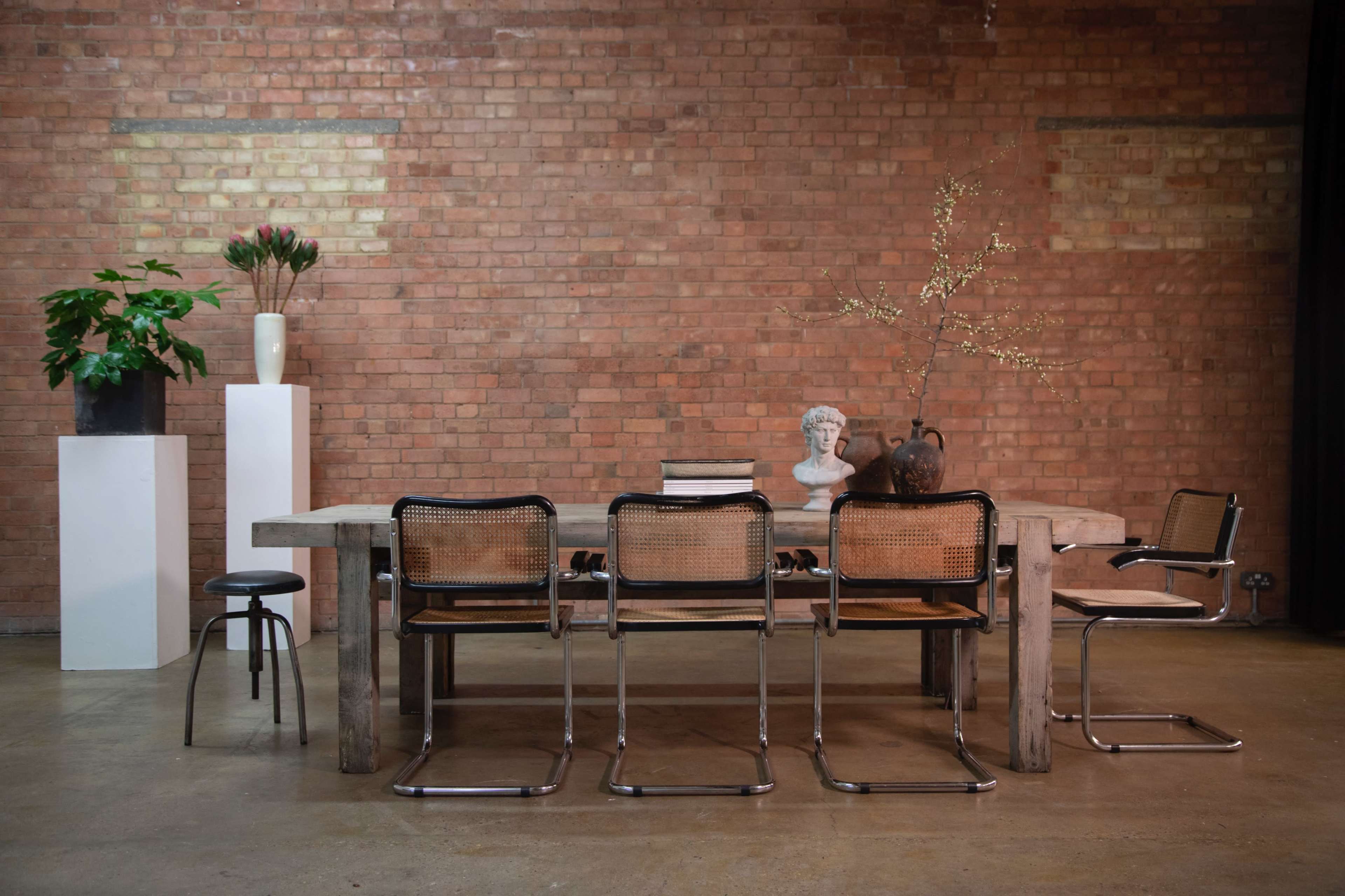 A minimalist dining area features a wooden table surrounded by several chairs, with a bust and decorative items on the table, set against a brick wall.