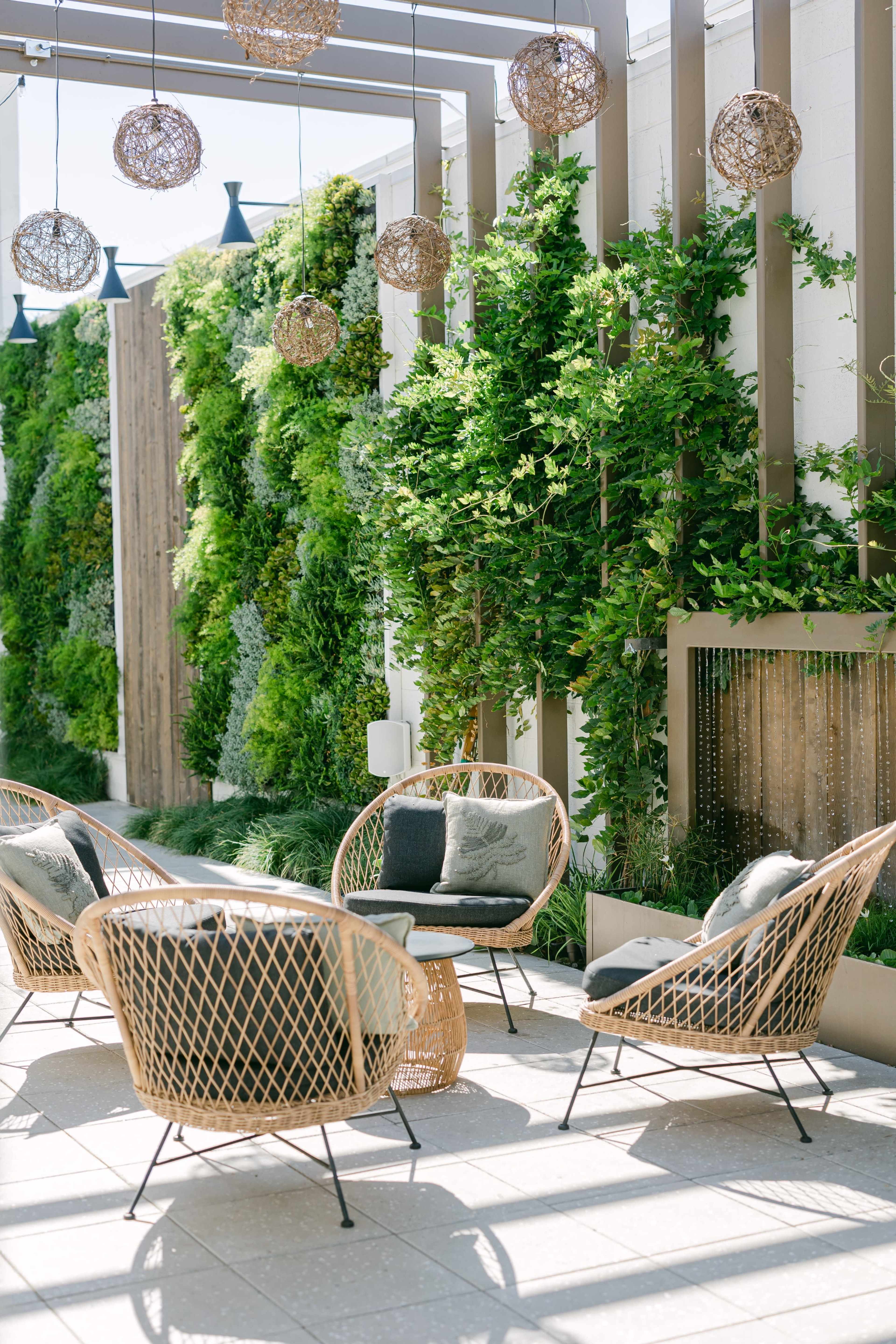 The scene features a set of four wicker chairs arranged around a small table in a greenery-filled outdoor space, with decorative hanging lights above.