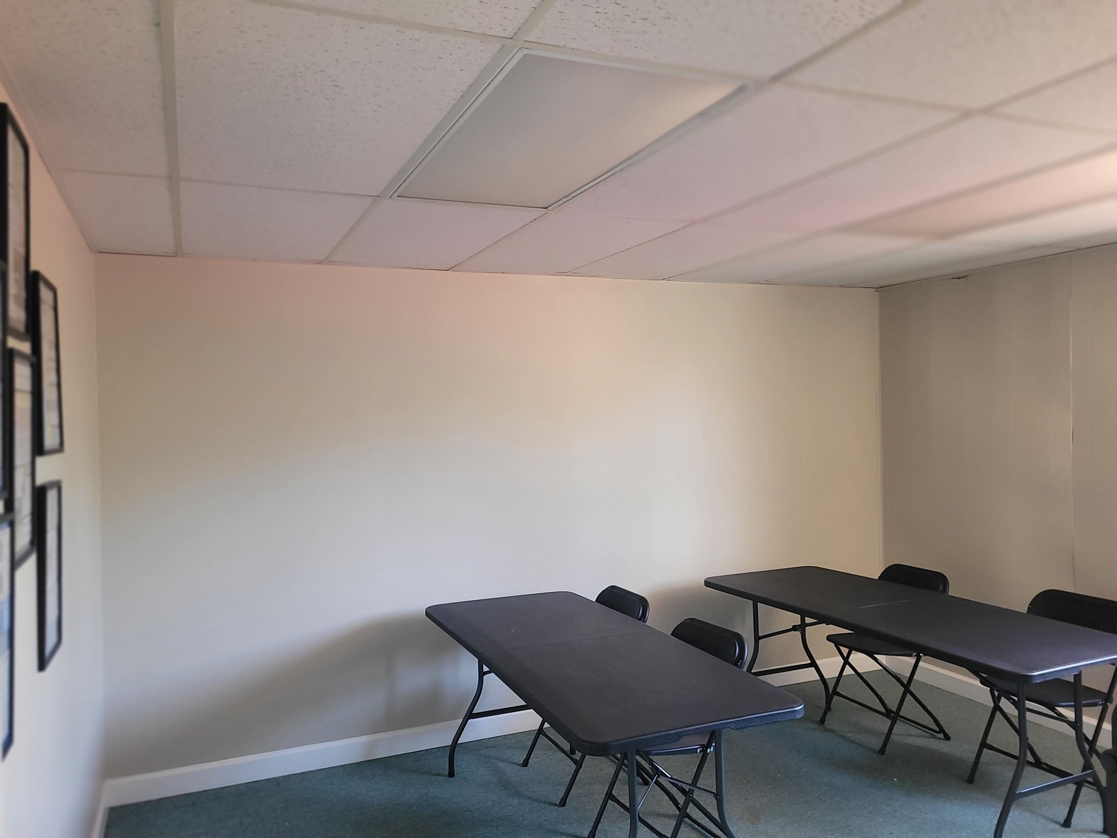 The image shows a sparsely furnished room with two black folding tables and several black chairs arranged in the space, against plain walls.