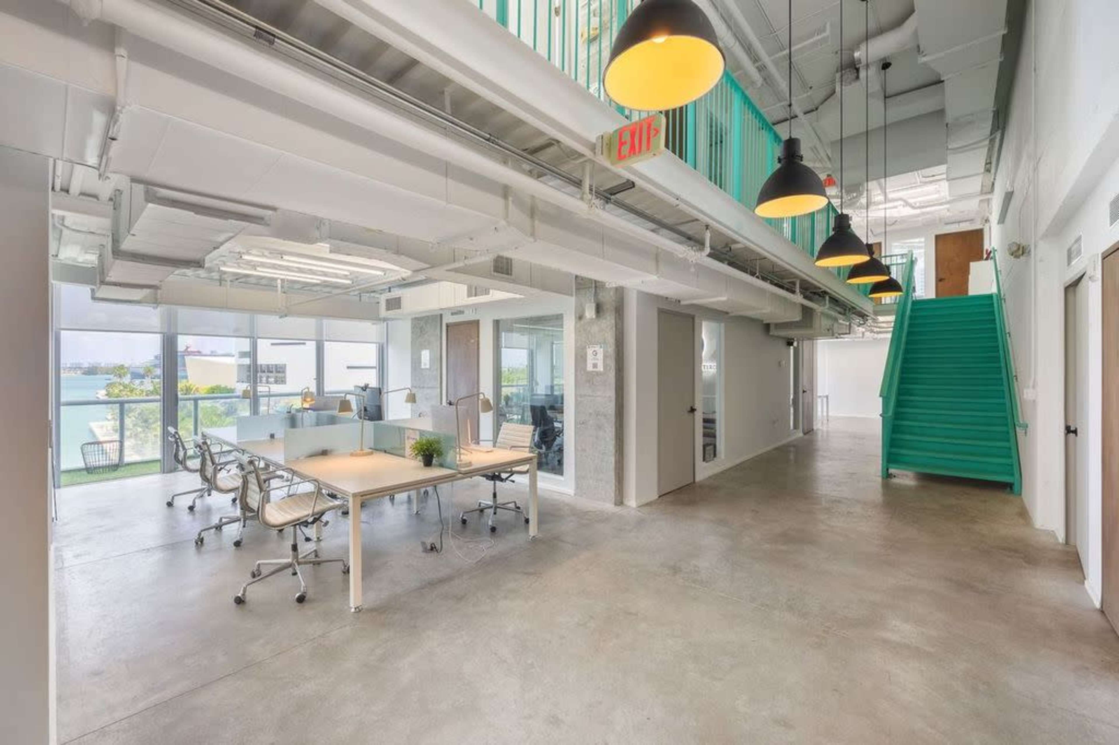 A modern office interior featuring a workspace with a large table and chairs, concrete flooring, and a turquoise staircase in the background.