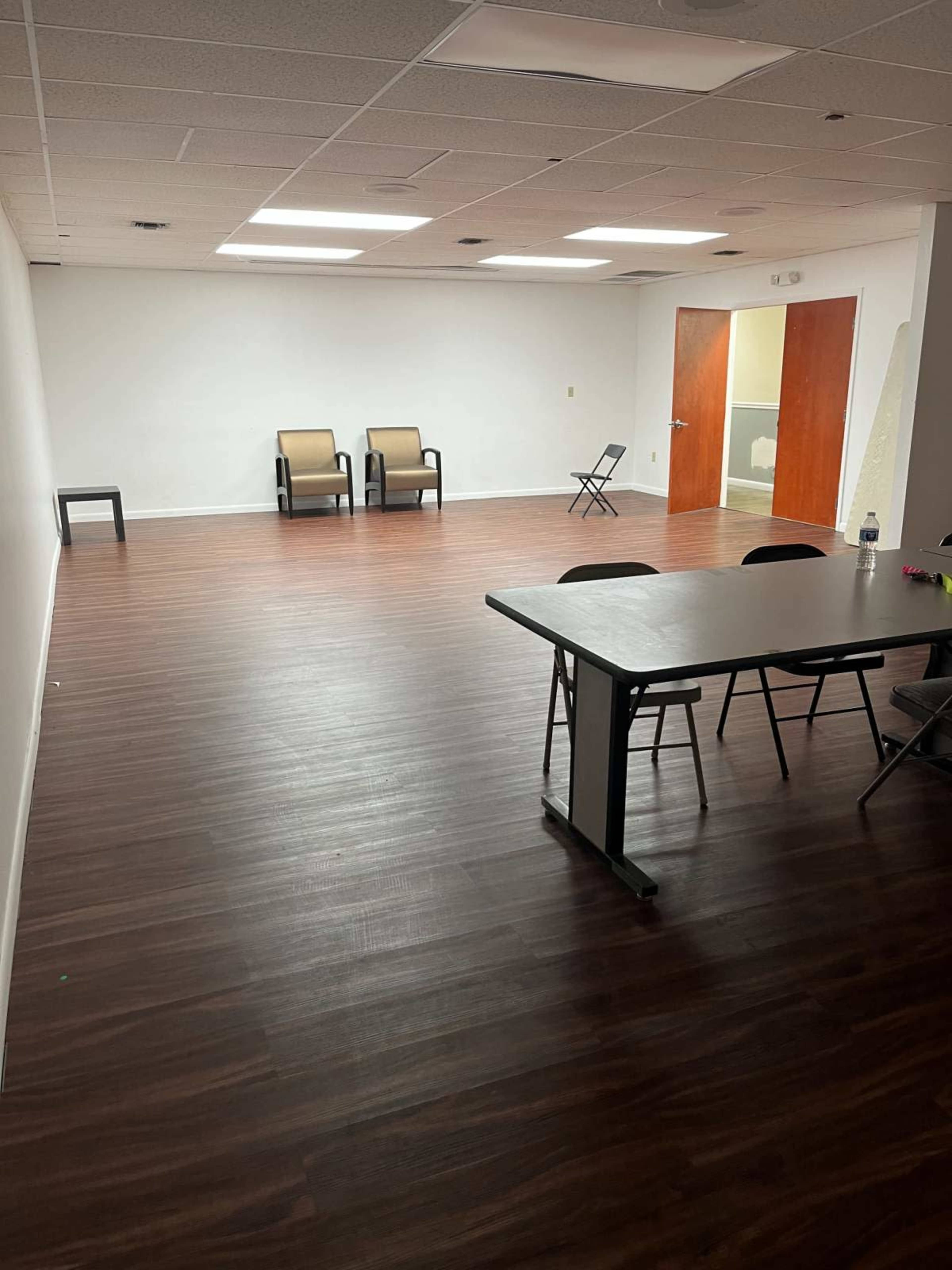 The image shows a spacious, empty room with brown wooden floors, two chairs against one wall, and a table with chairs set up in the foreground.