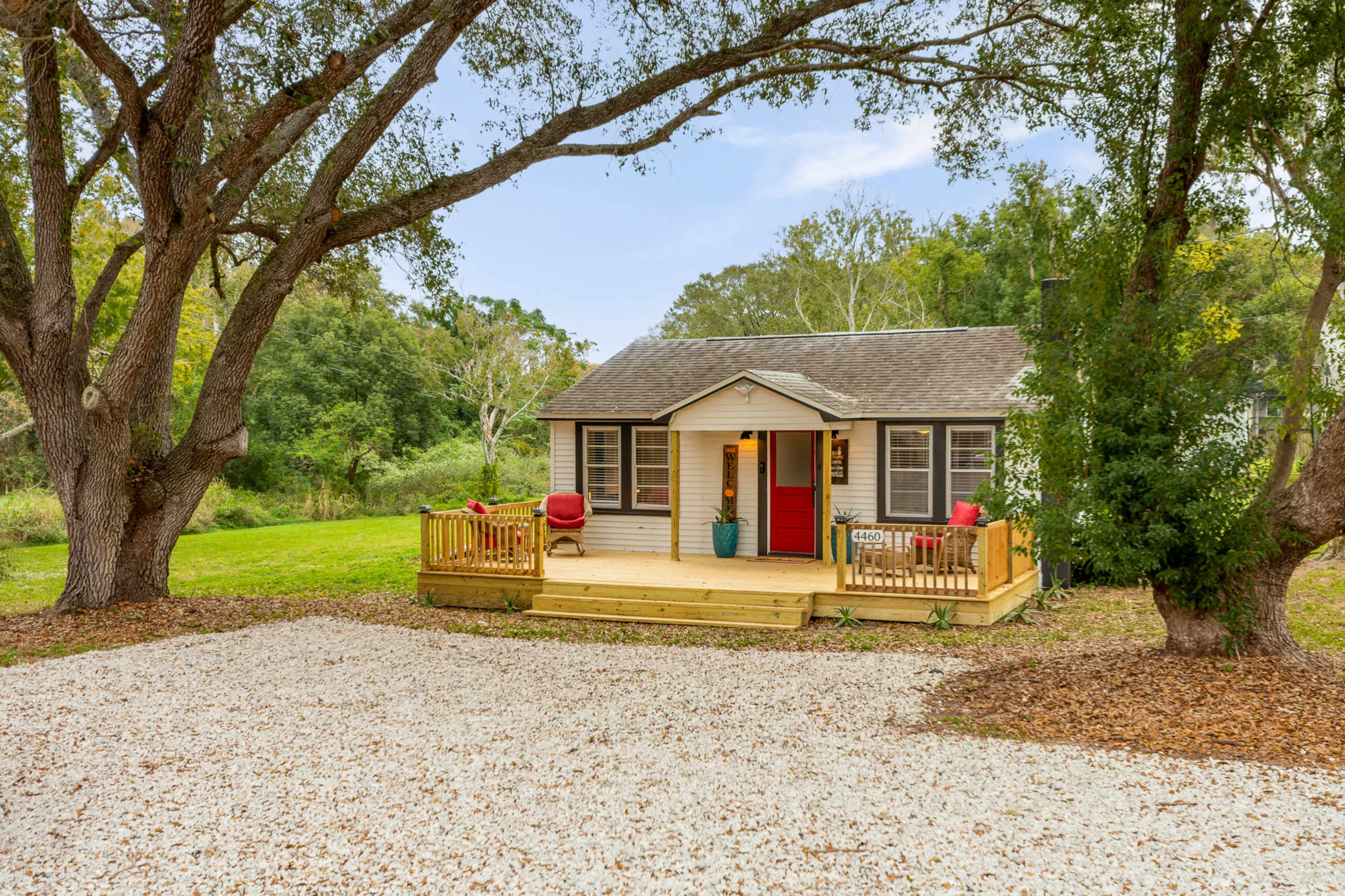A small, single-story house with a red door and a front porch is situated in a grassy area surrounded by trees.