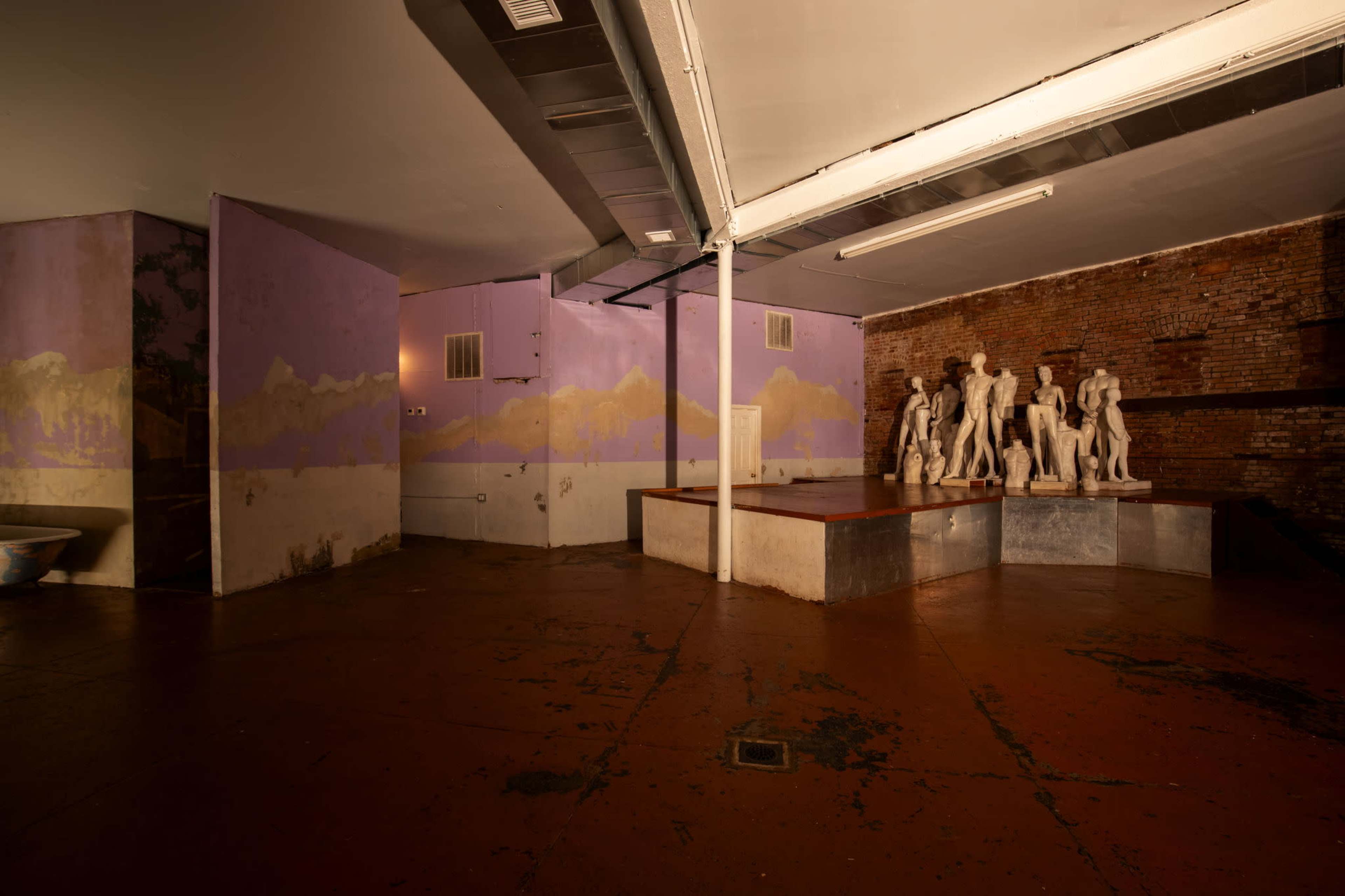 The image shows an empty room with brick walls, a painted mural of mountains, and a platform displaying several white sculptures.