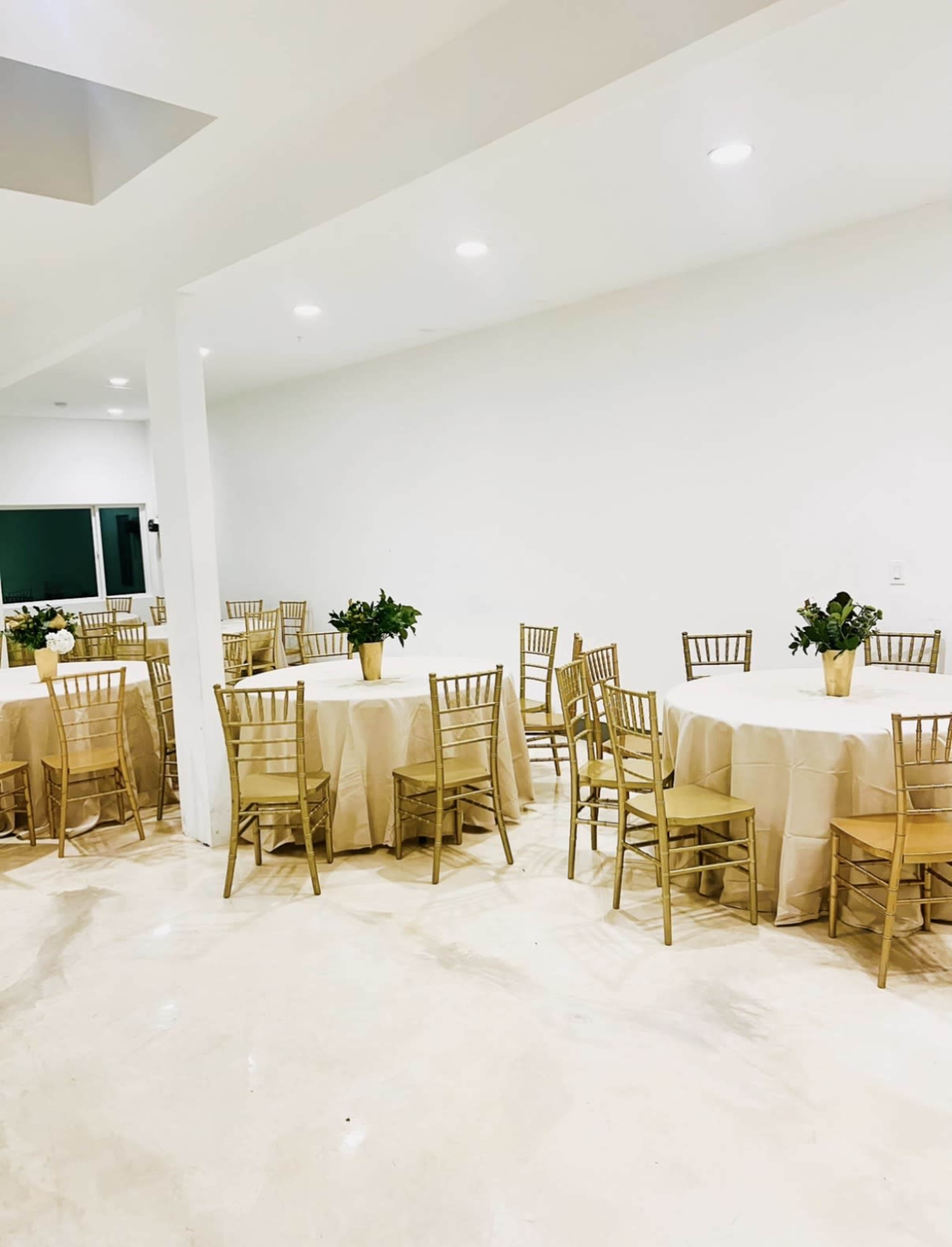 The image shows a modern event space with several round tables covered in beige tablecloths and surrounded by gold Chiavari chairs, set against a plain white wall.