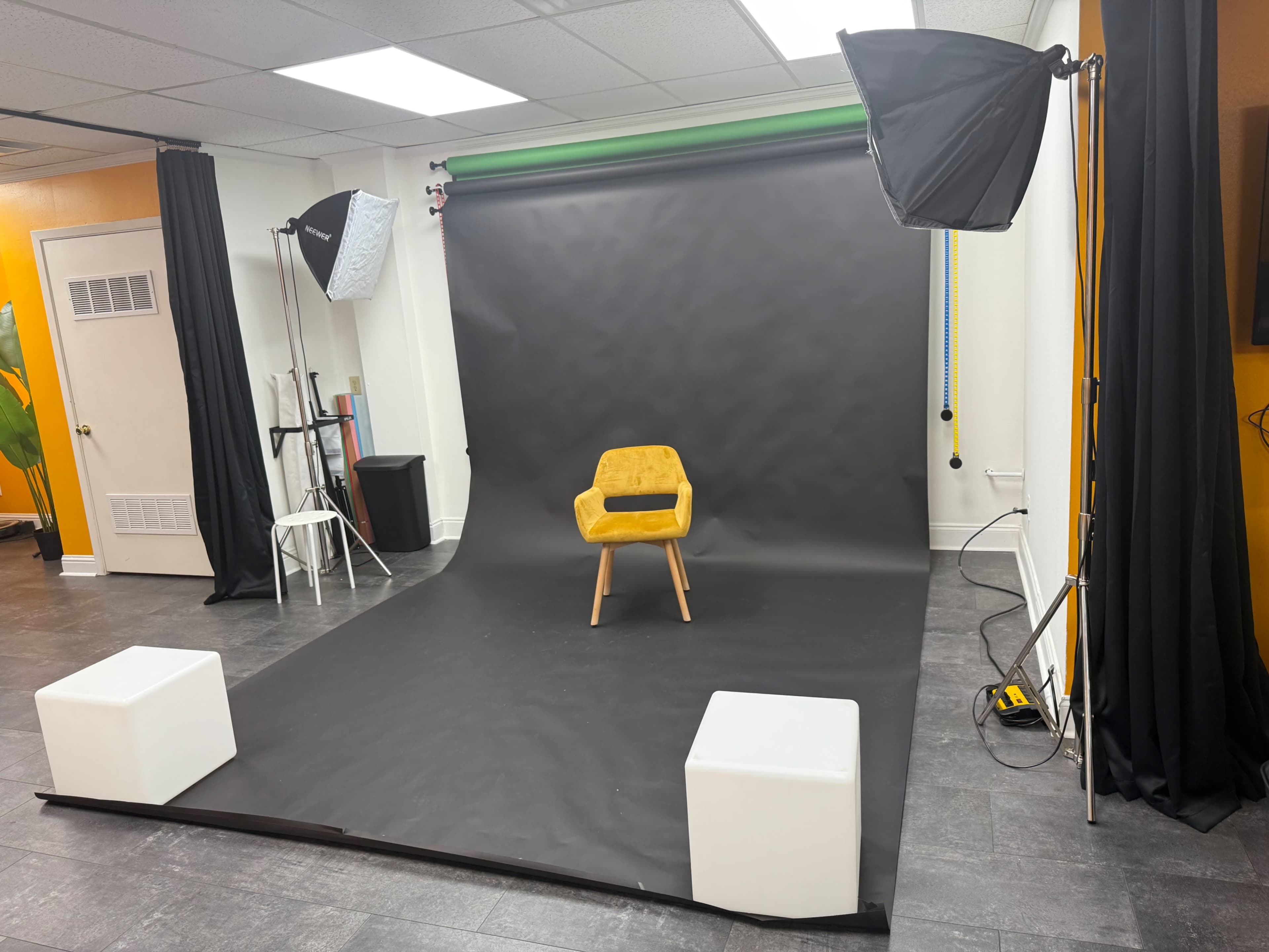 The image shows a photography studio with a black backdrop, a yellow chair in the center, and lighting equipment on either side.