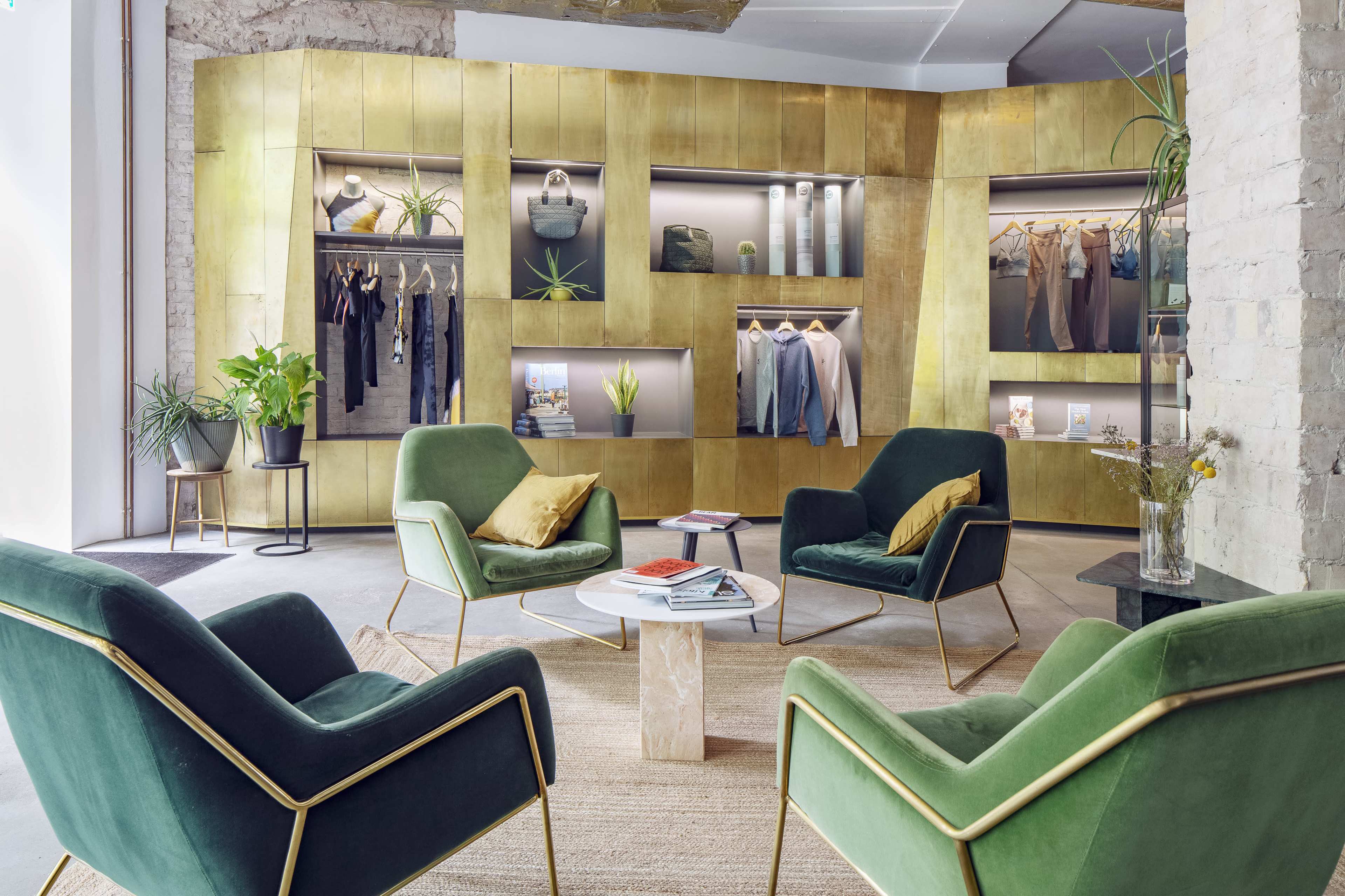 A modern interior with gold-paneled walls, green velvet chairs, a white circular table, and various clothing displayed on racks.