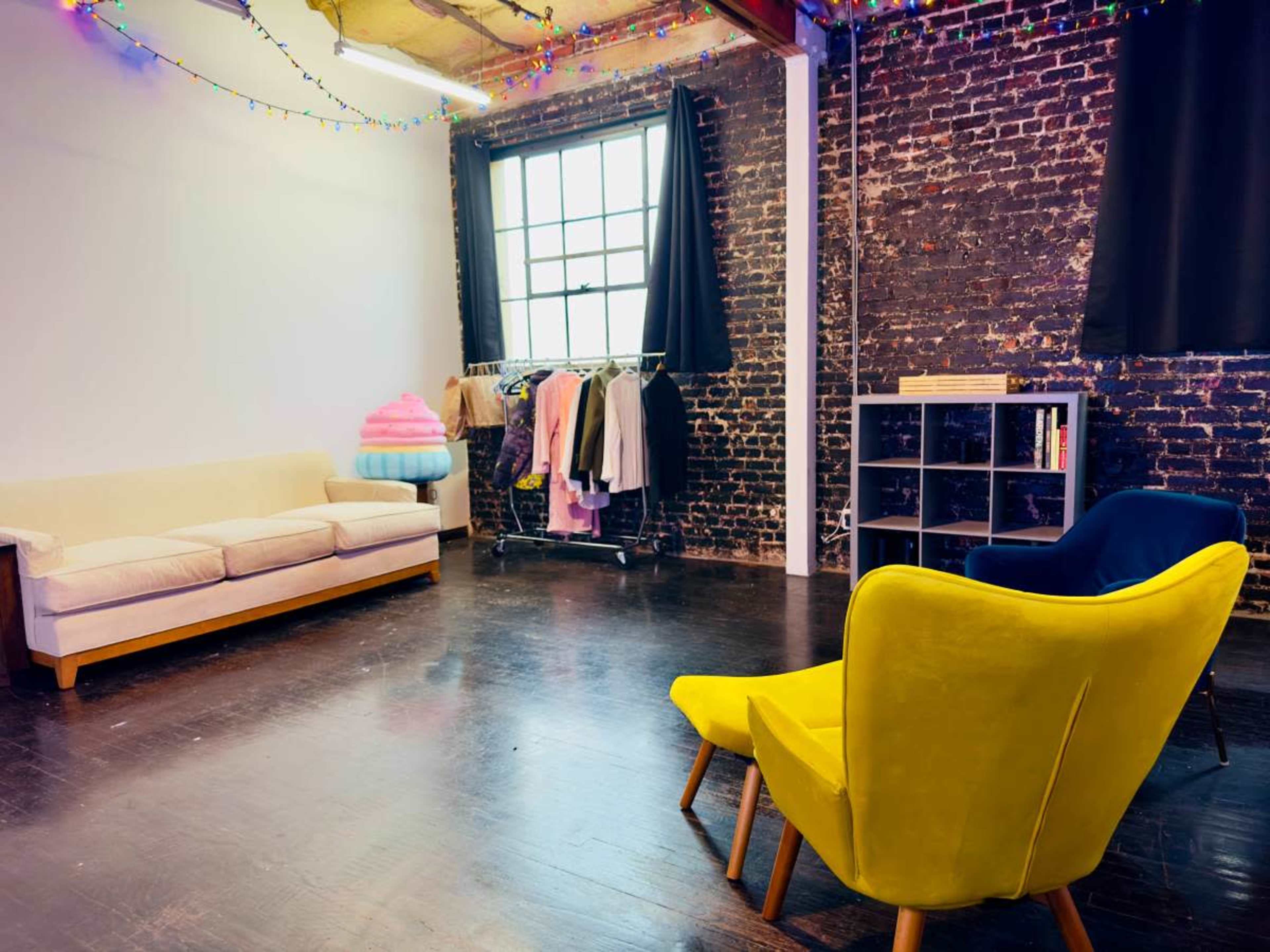 A minimalist room with exposed brick walls, a beige couch, two brightly colored chairs, a clothing rack, and a bookshelf.