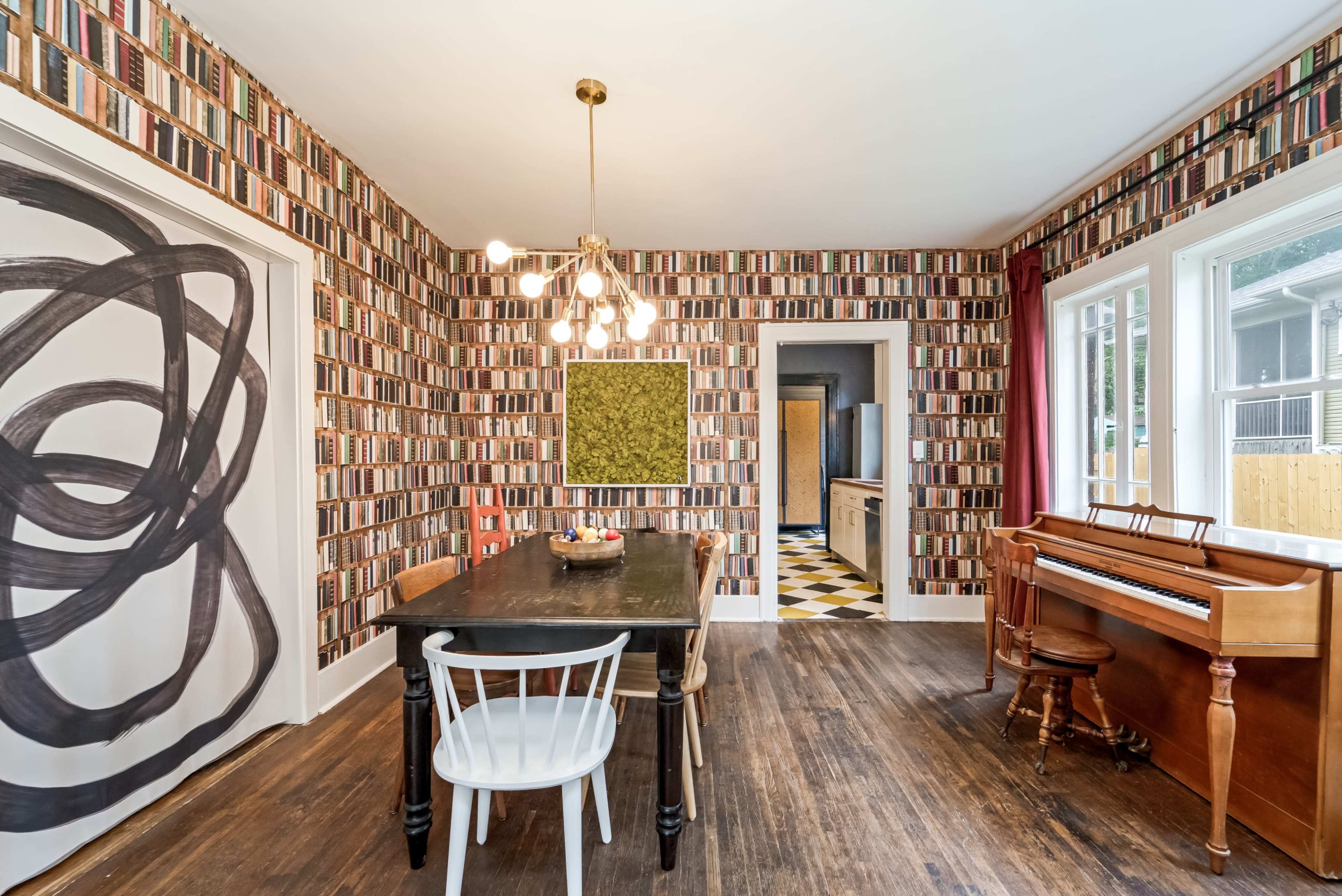 A dining room features a table with chairs, a piano, and walls decorated with a book-patterned wallpaper.