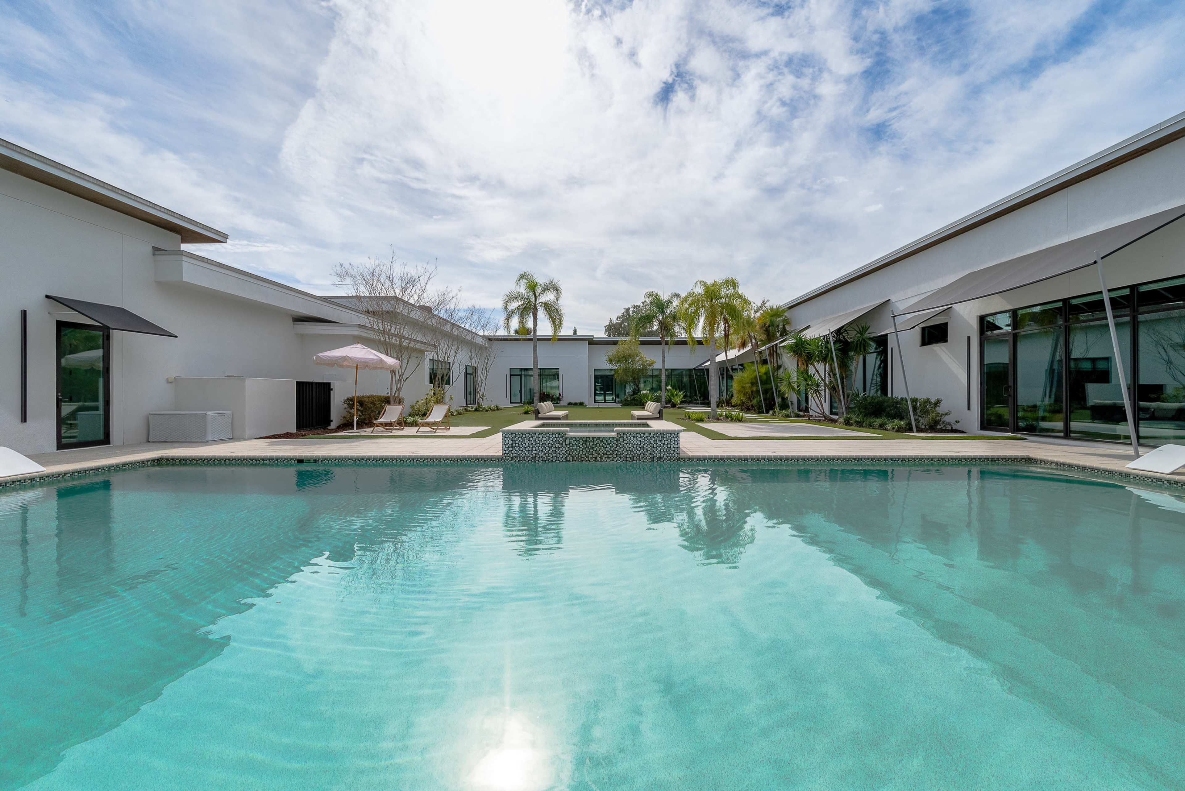 The image shows a modern house with a pool in the foreground, surrounded by palm trees and landscaped gardens.