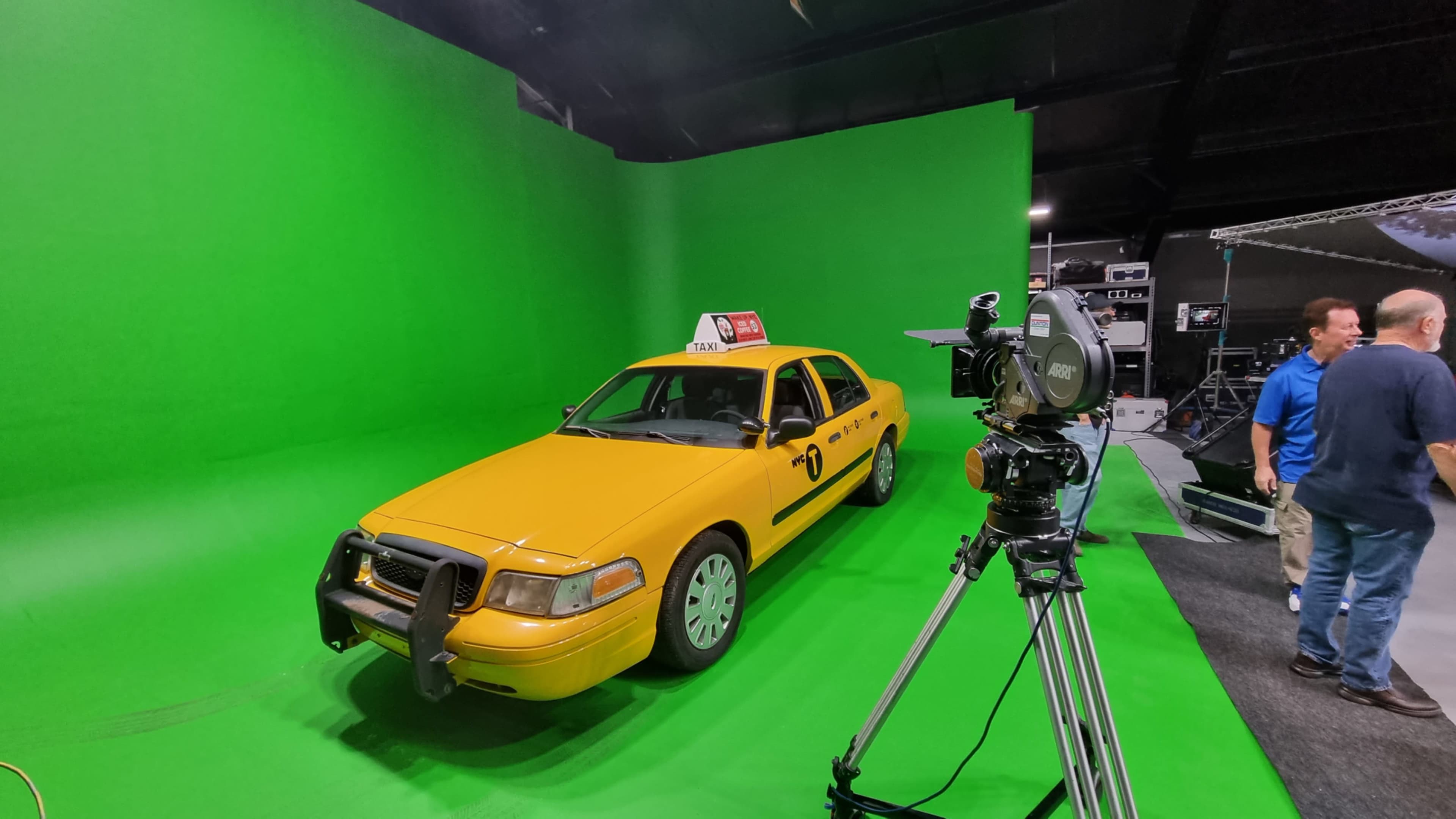 A yellow taxi cab is positioned in front of a green screen in a studio setting, with a camera setup nearby.