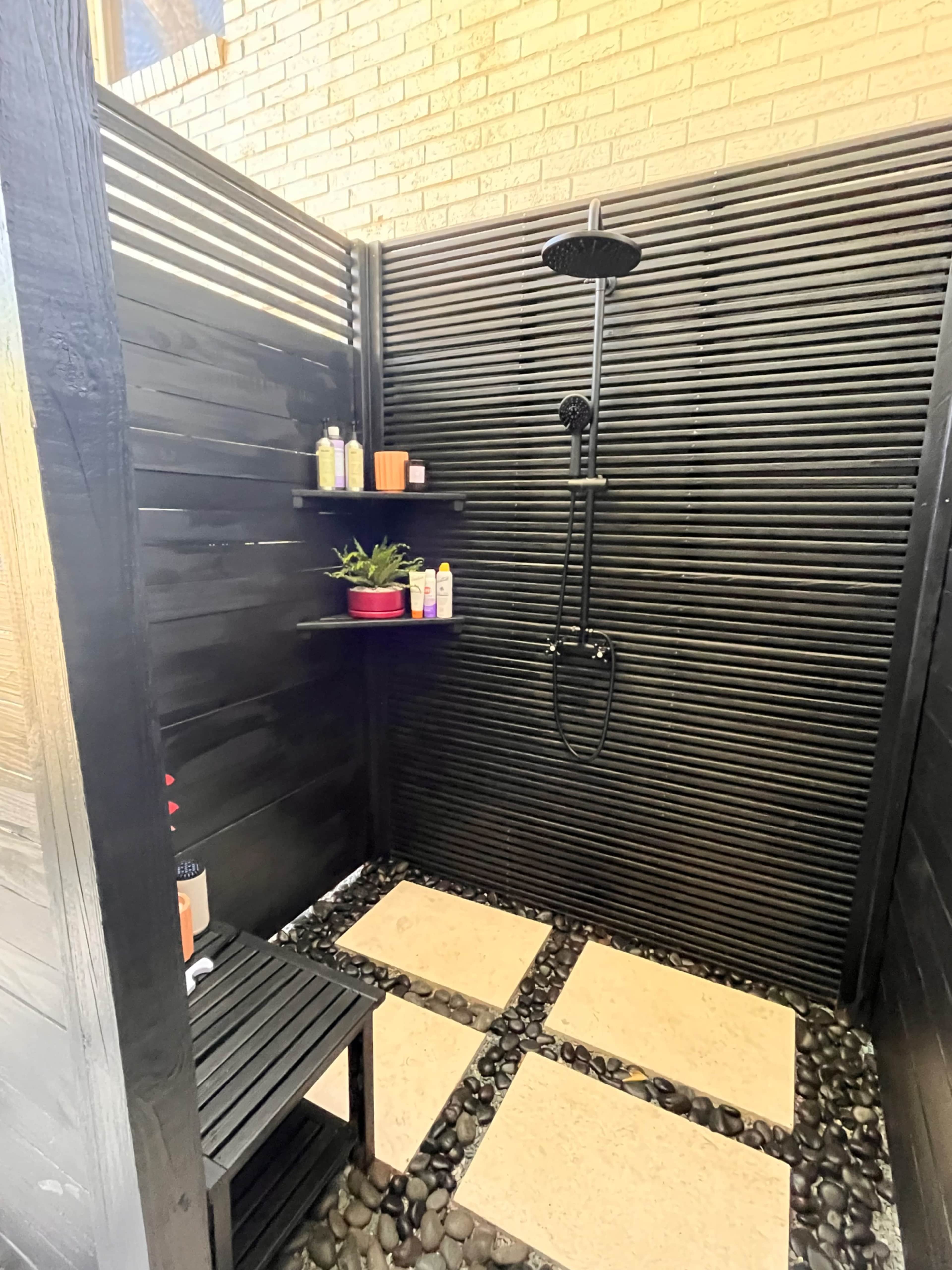 An outdoor shower area with black wooden slats, a showerhead, shelves for toiletries, a small planter, and stone flooring.
