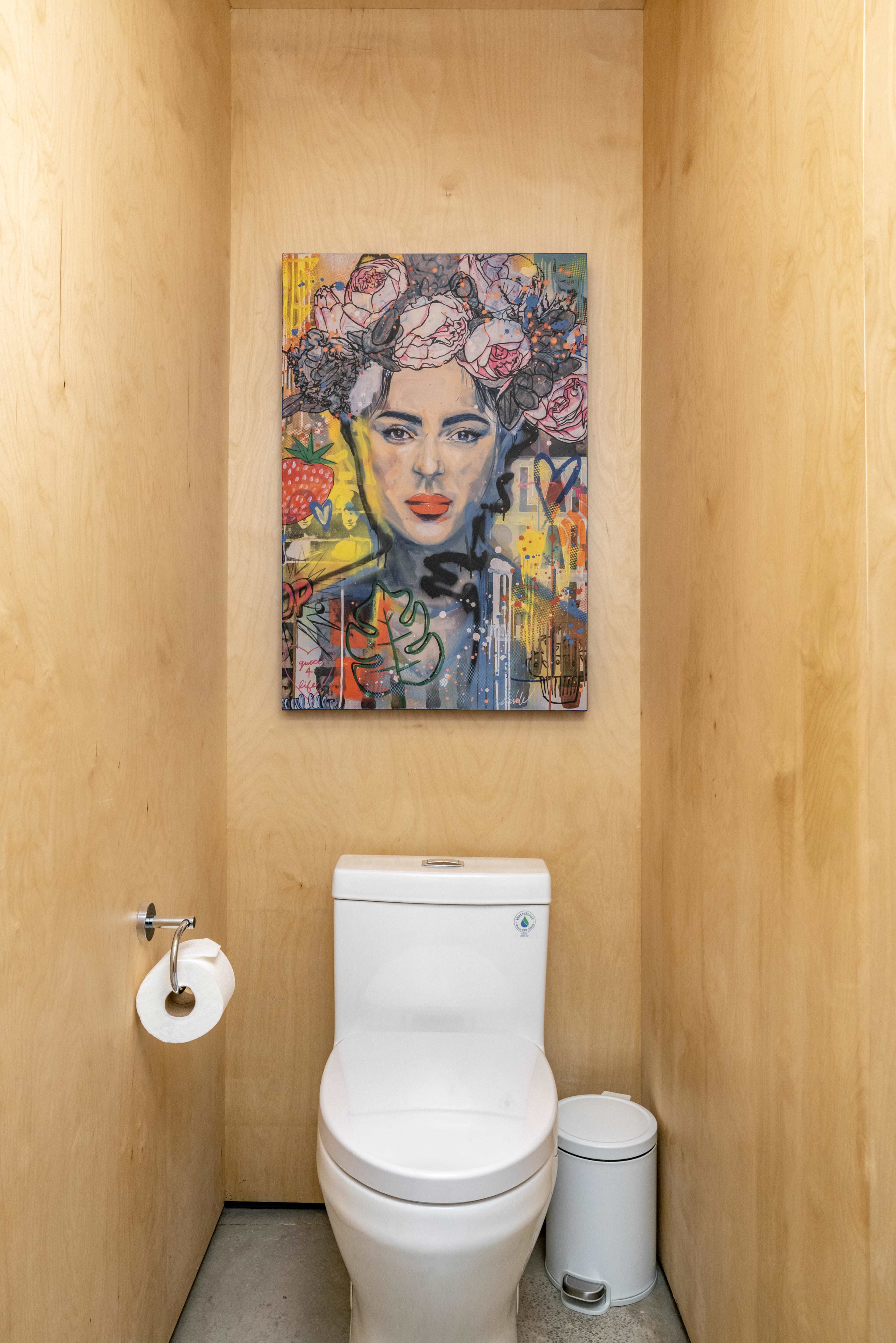 A bathroom stall with a toilet, a roll of toilet paper, a small trash can, and a vibrant portrait on the wall.