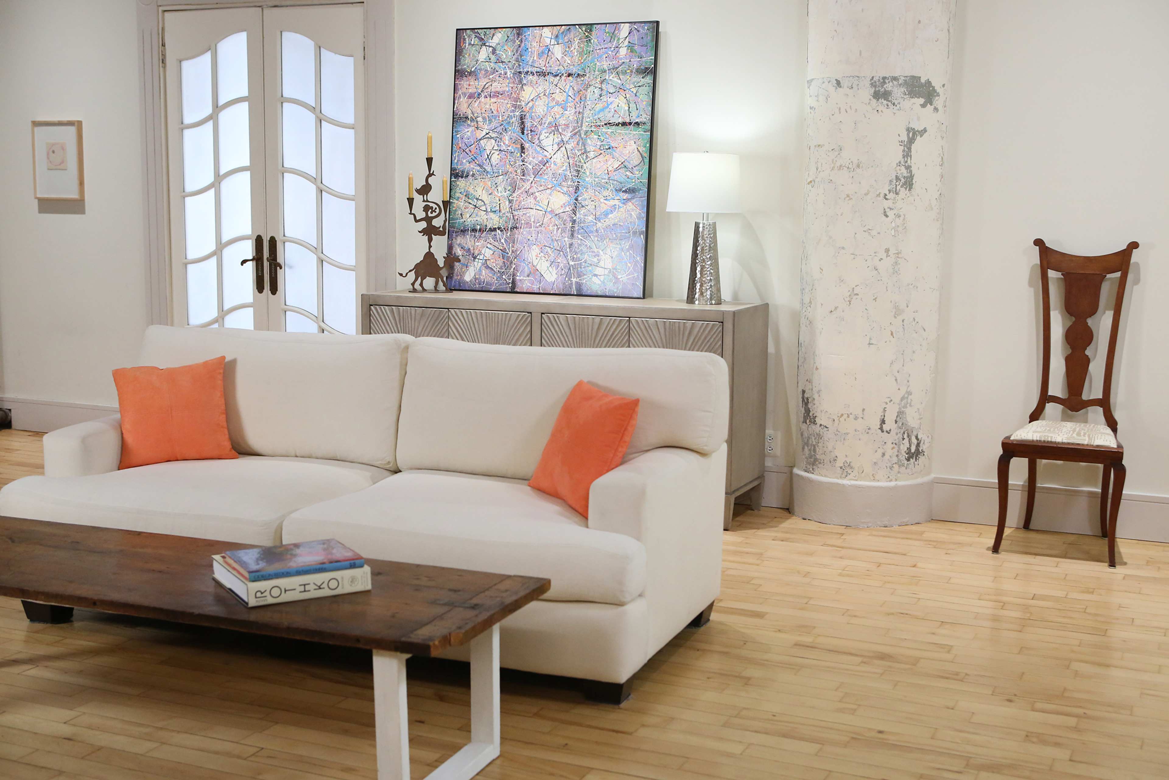 A white sofa with orange cushions is positioned in a minimalist living room featuring a wooden coffee table, a light-colored cabinet, a contemporary lamp, and a single wooden chair against a textured wall.