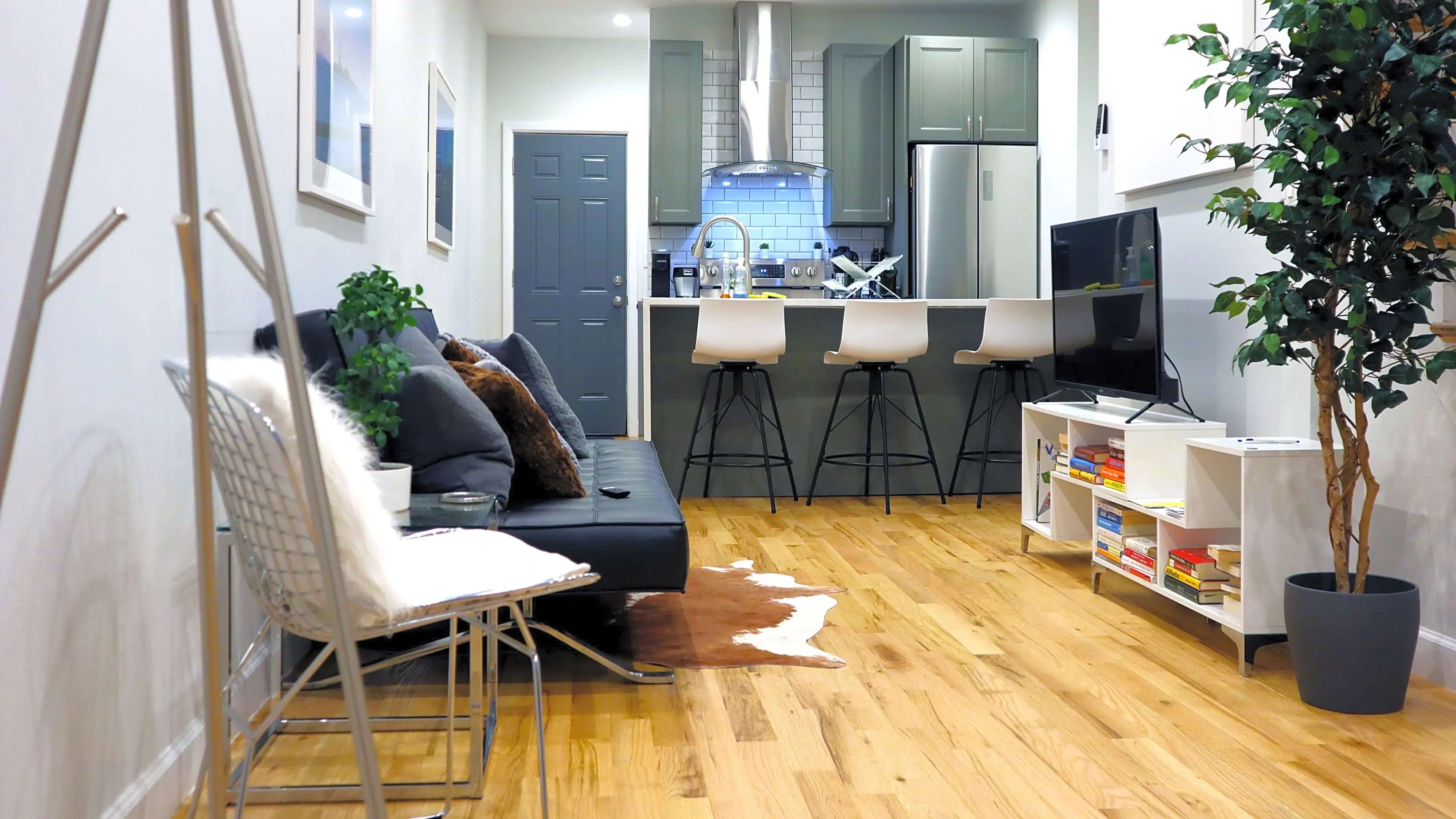 The image shows a modern living area with a black sofa, a small coffee table, and a kitchenette in the background featuring bar stools and a plant.