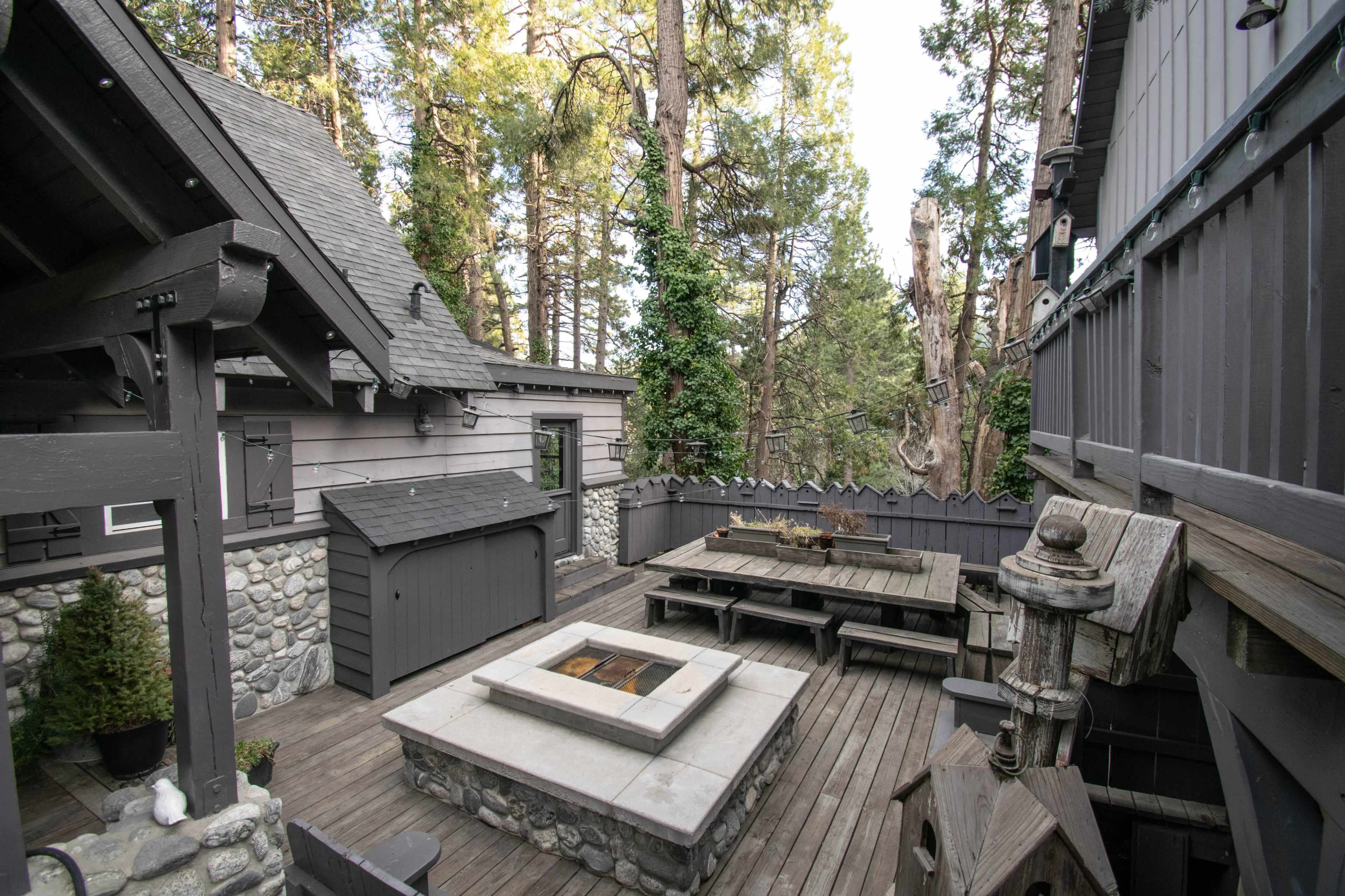 A wooden deck area adjacent to a house, featuring a stone fire pit and a dining table surrounded by tall trees.