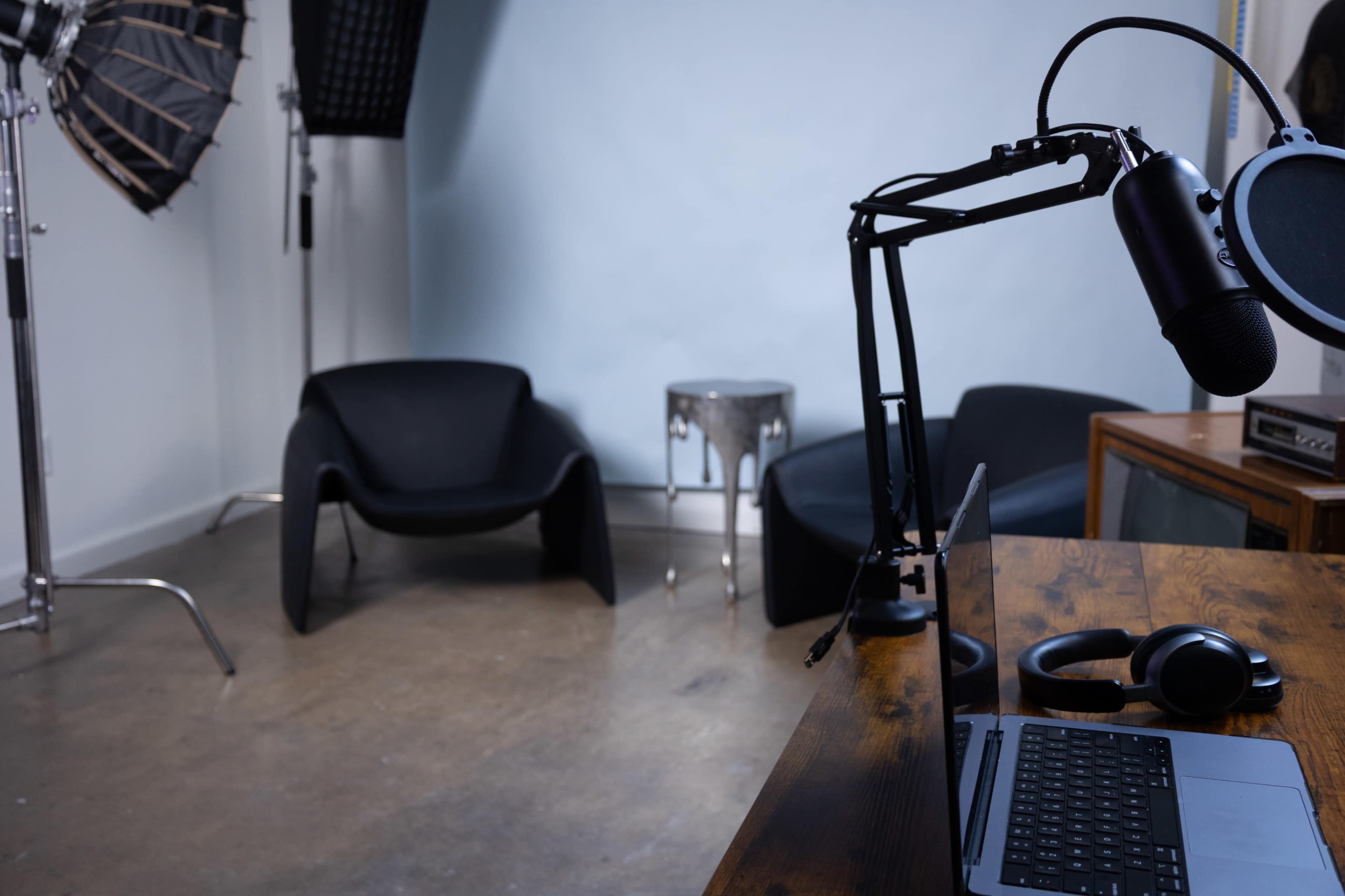 The image shows a minimalist studio setup with a laptop on a wooden table, a microphone mounted on an arm, and two black chairs against a light blue wall.