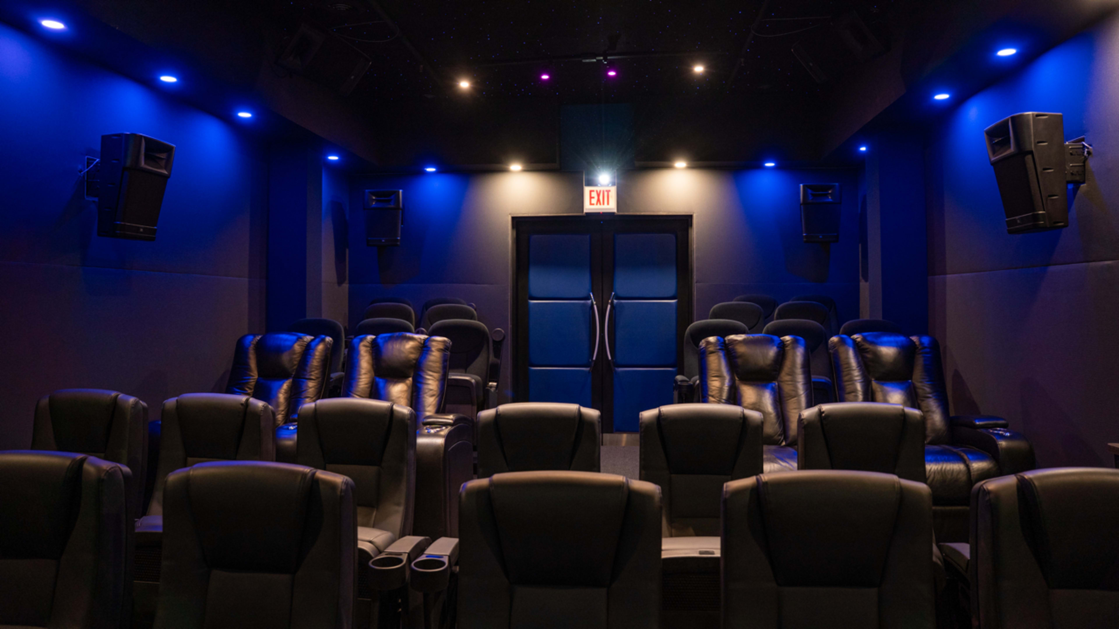 The image shows a dark, modern home theater with plush black reclining seats arranged in rows, illuminated by blue LED lights.