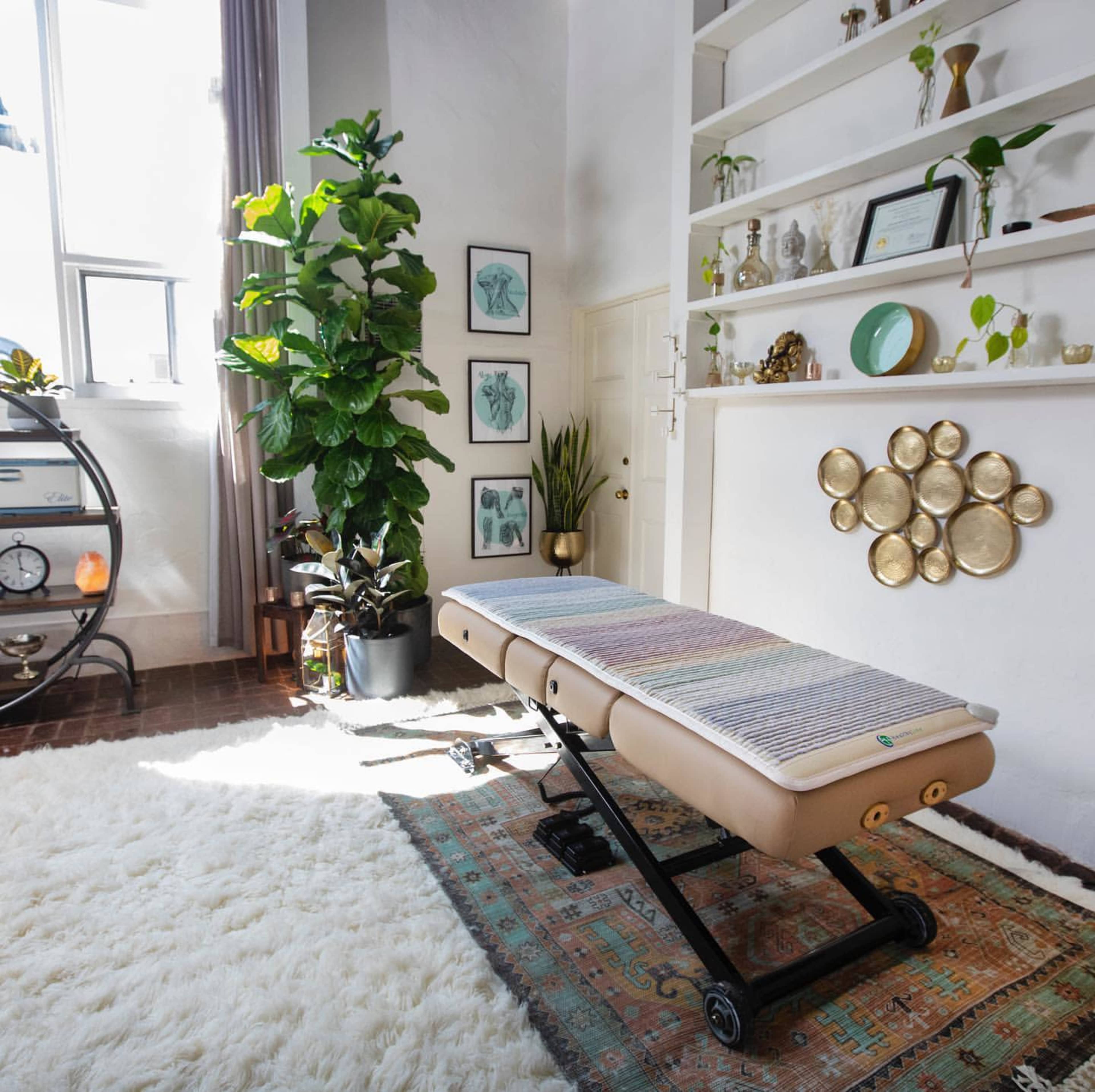 A massage table positioned in a well-lit room with decorative plants and framed artwork on the walls.