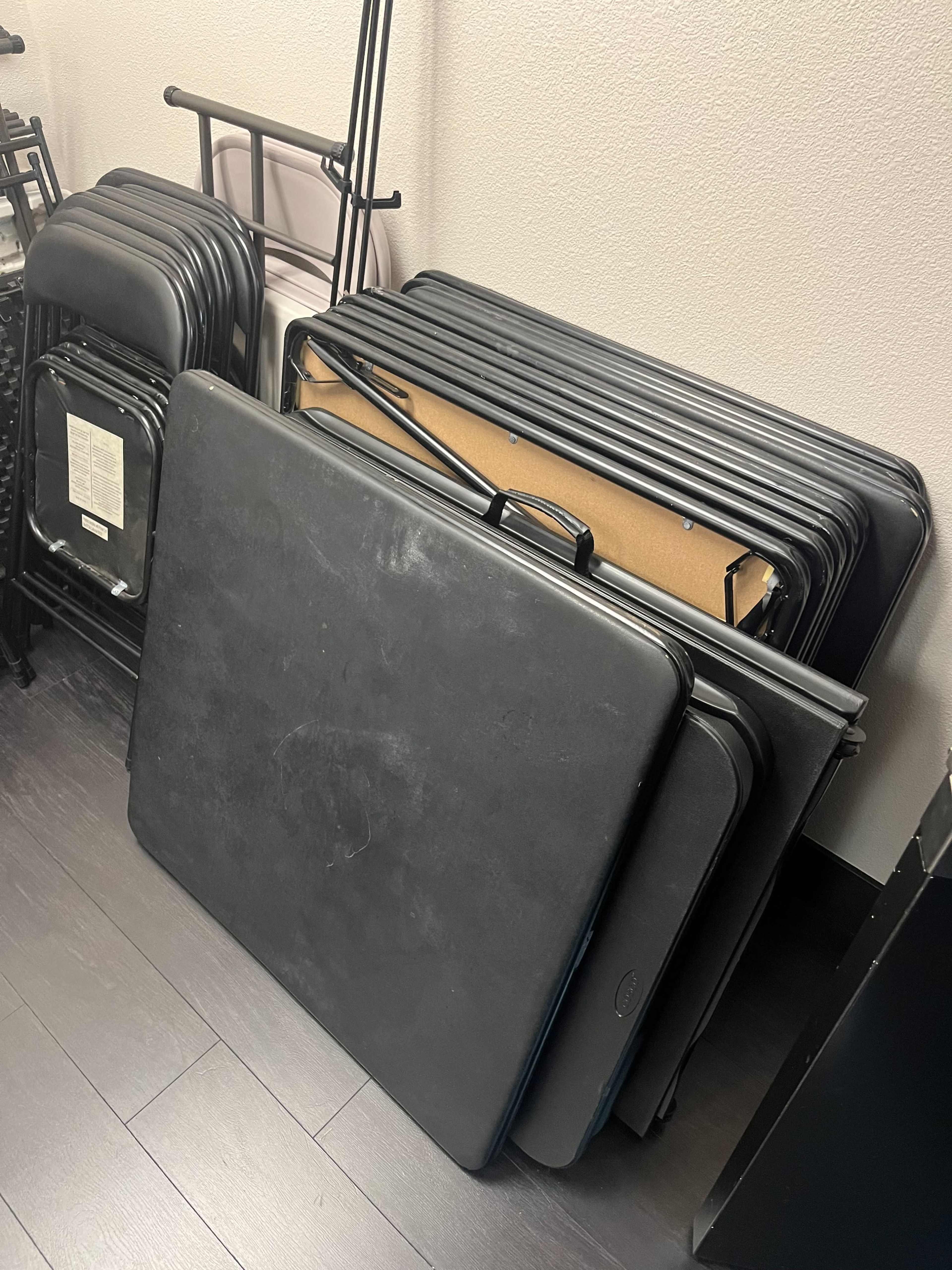 A stack of black folding tables is leaned against a wall in a storage area.