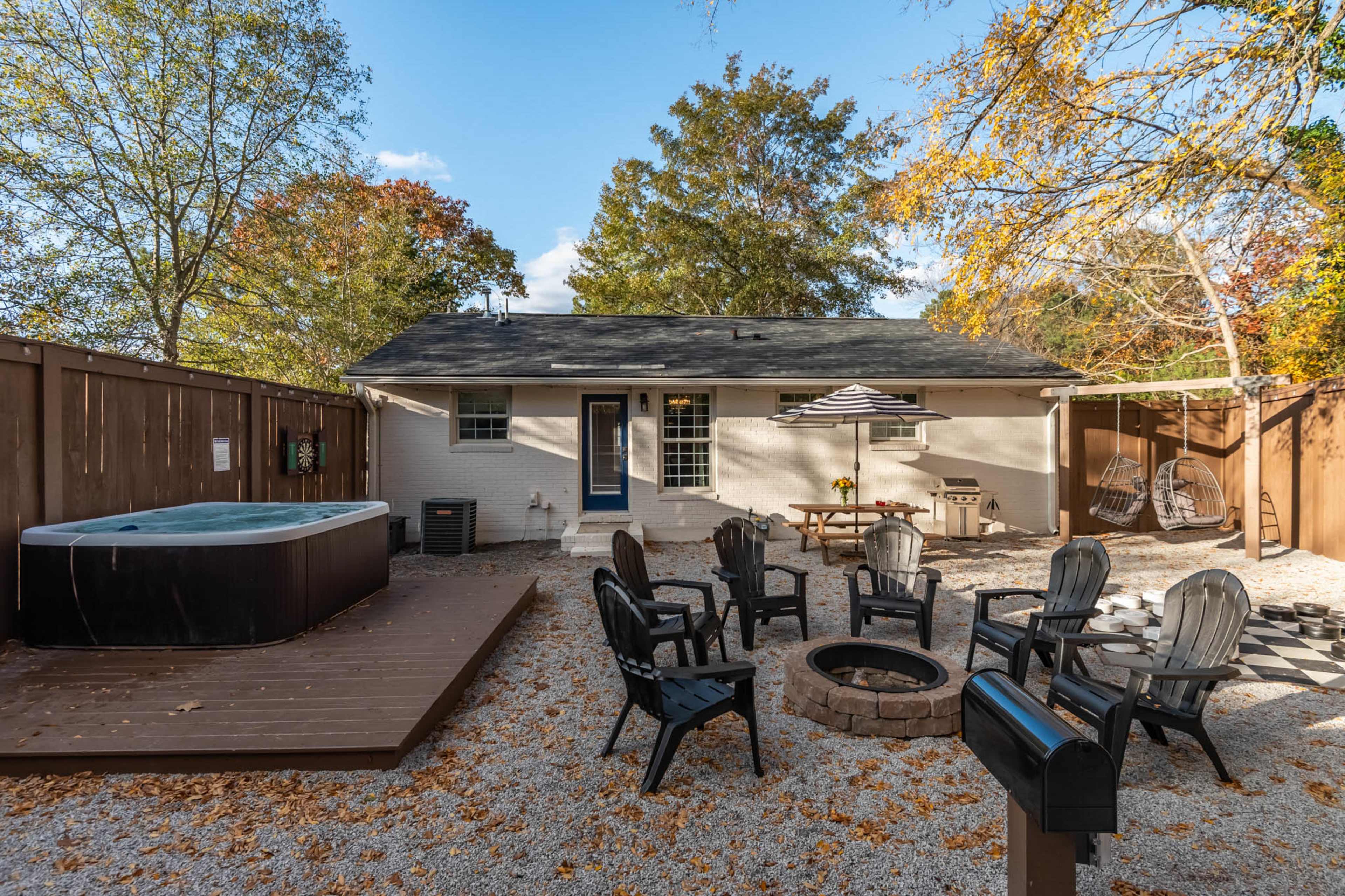 The image shows a backyard area with a hot tub, fire pit surrounded by chairs, and a patio space, all enclosed by wooden fencing and featuring autumn foliage.