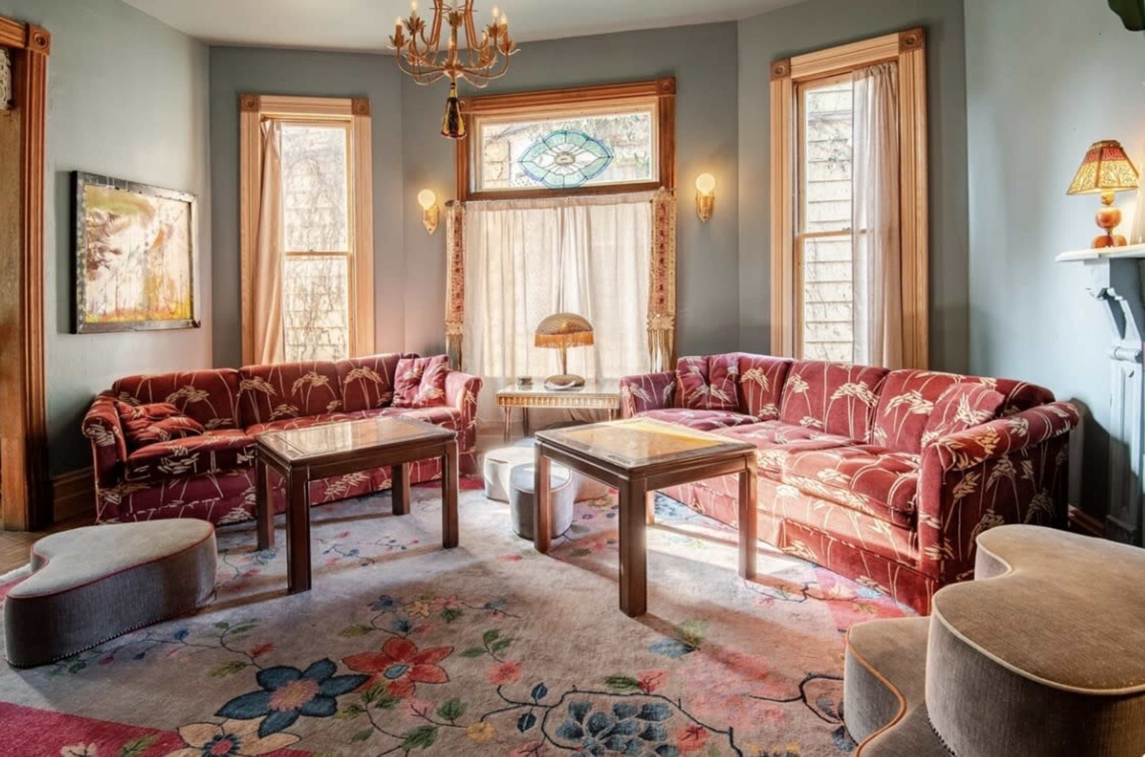 The image shows a living room with two red-patterned couches, a coffee table, and large windows adorned with sheer curtains, illuminated by natural light.
