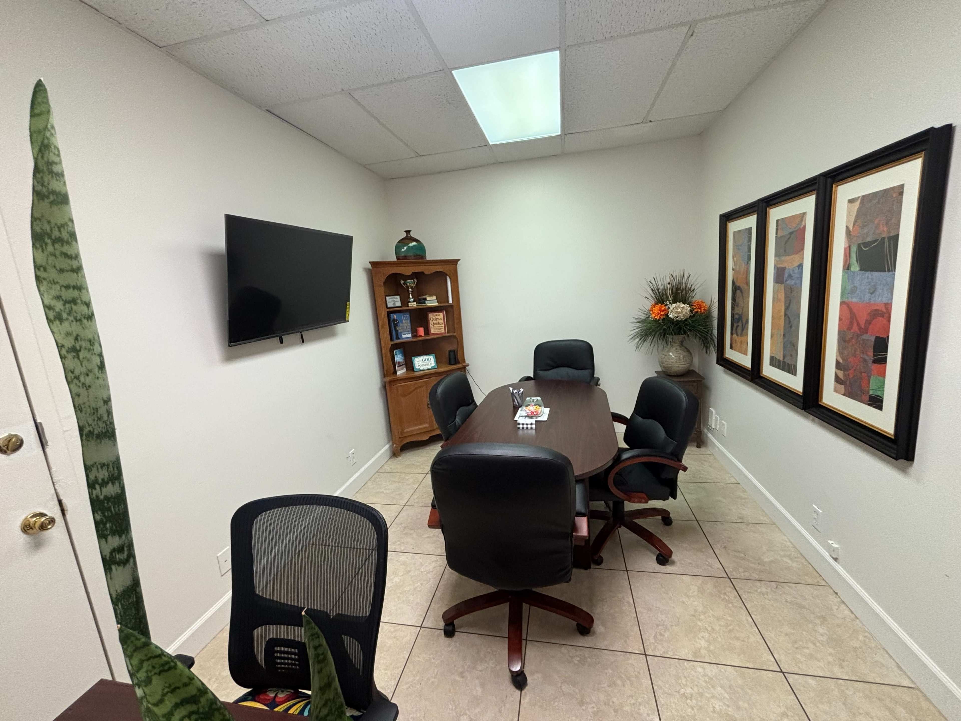 The image shows a small conference room with a rectangular table surrounded by four black chairs, a television on the wall, a wooden cabinet, and framed artwork.
