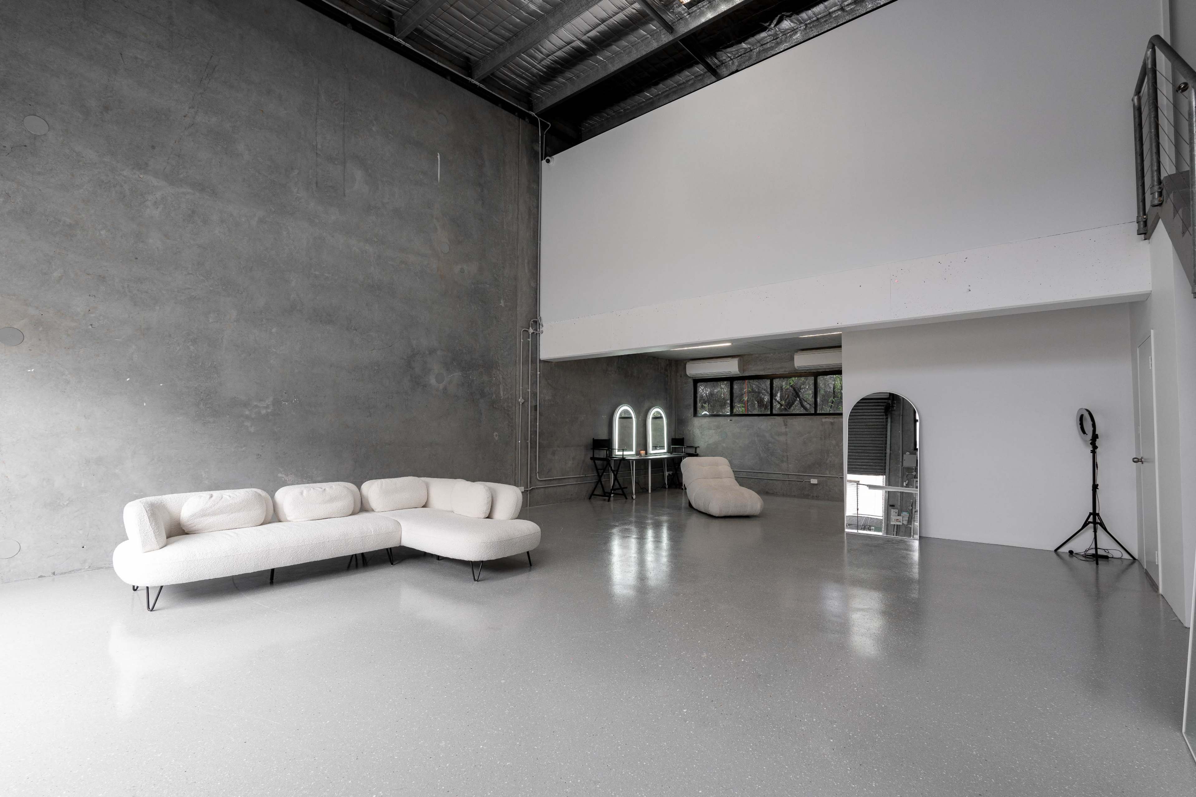 A spacious, minimalist room with a gray concrete wall, a light-colored sofa, and a large mirror, accompanied by two vanity mirrors and a sleek chair in the background.