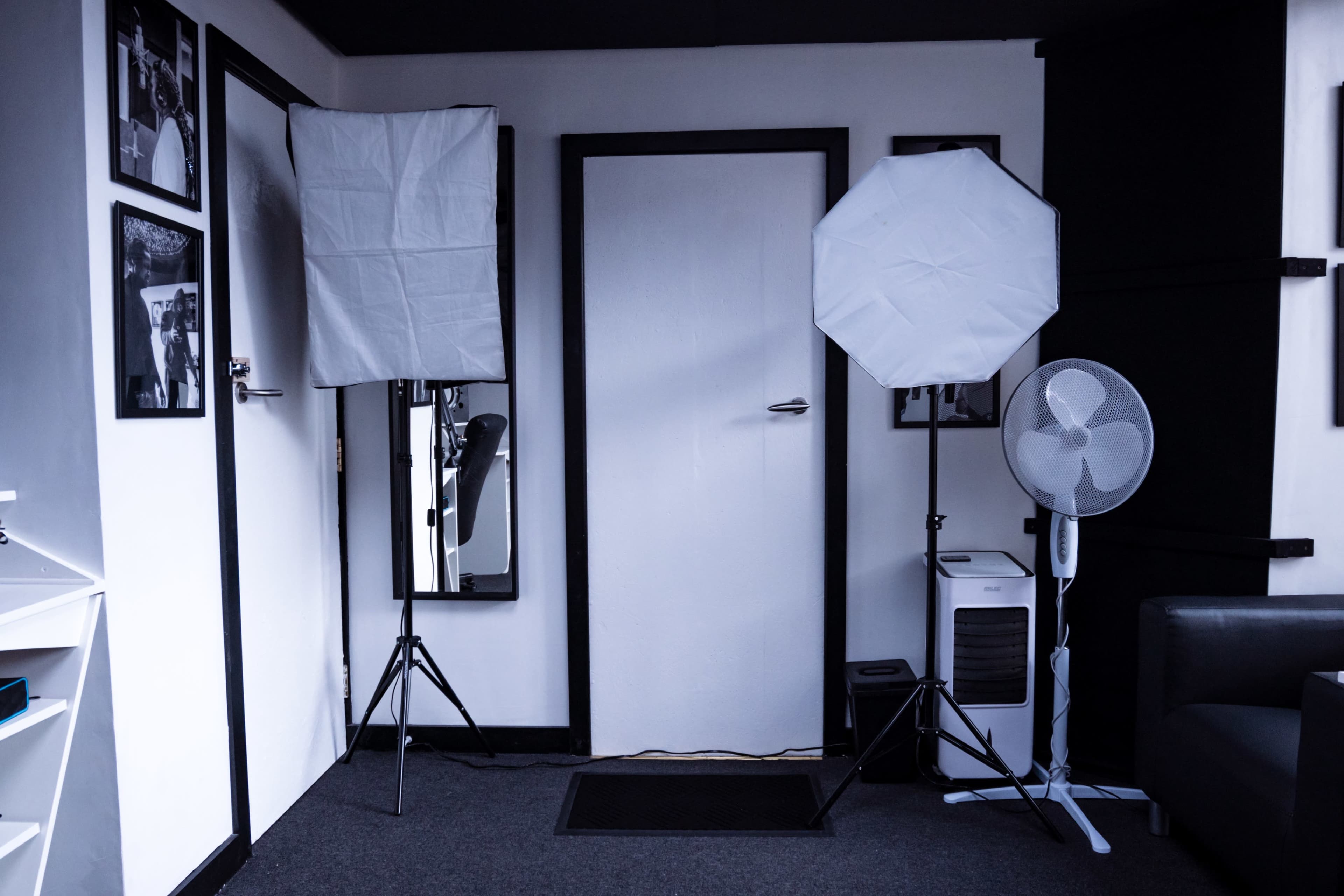 A room with two softbox lights on stands, a door, a fan, and a small air conditioning unit, alongside framed photographs on the walls.