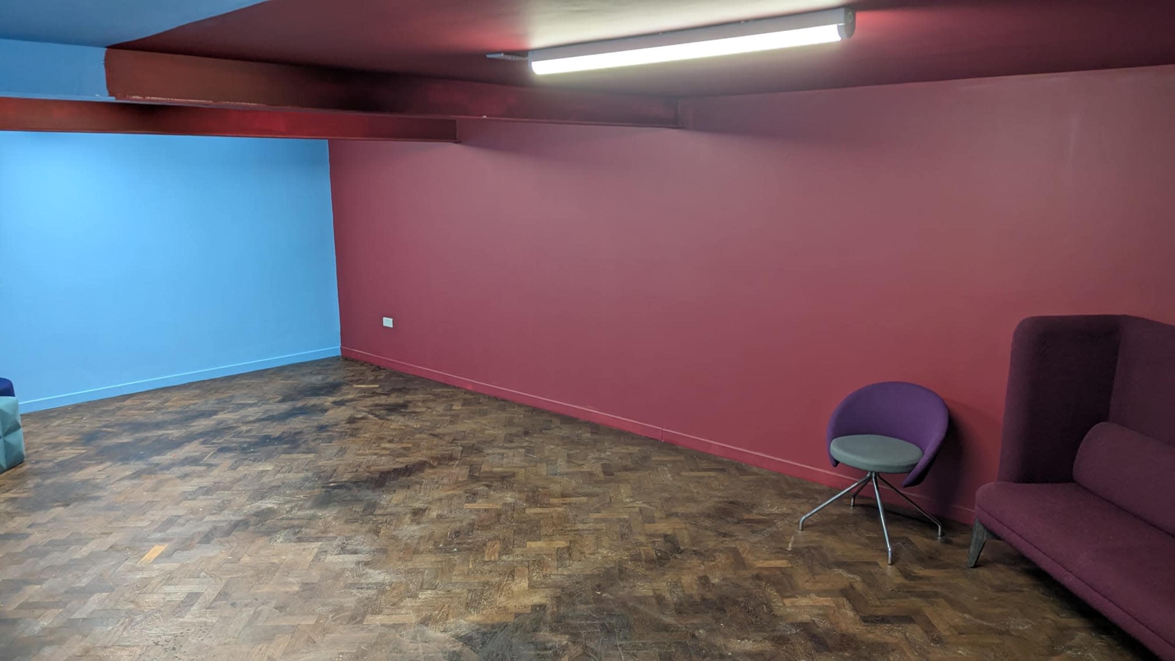 The image shows a room with two colored walls—one blue and one maroon—and a wooden floor, featuring a chair and a couch in the corner.