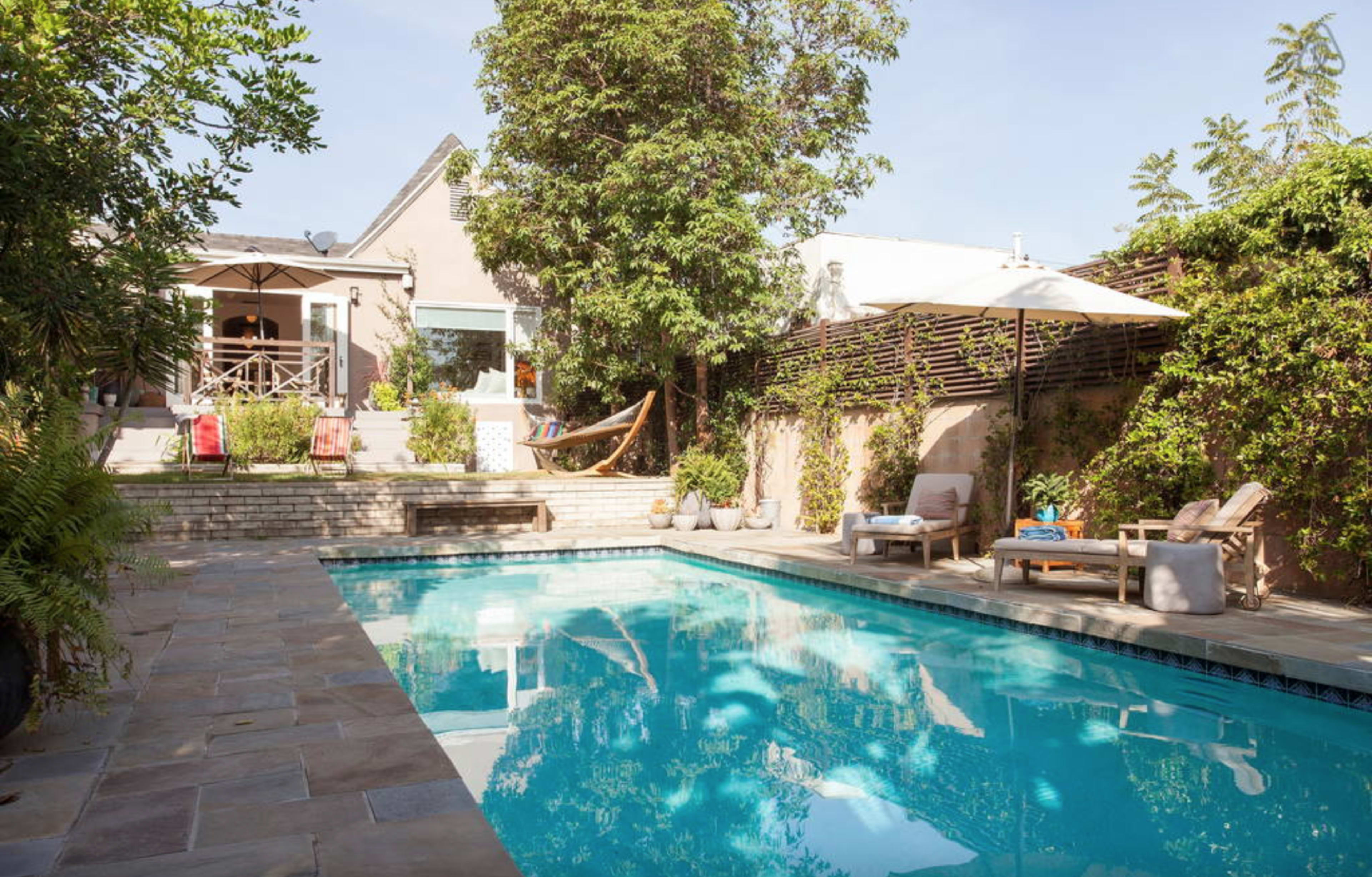 The image shows a backyard with a swimming pool surrounded by lounge chairs, plants, and a house in the background.
