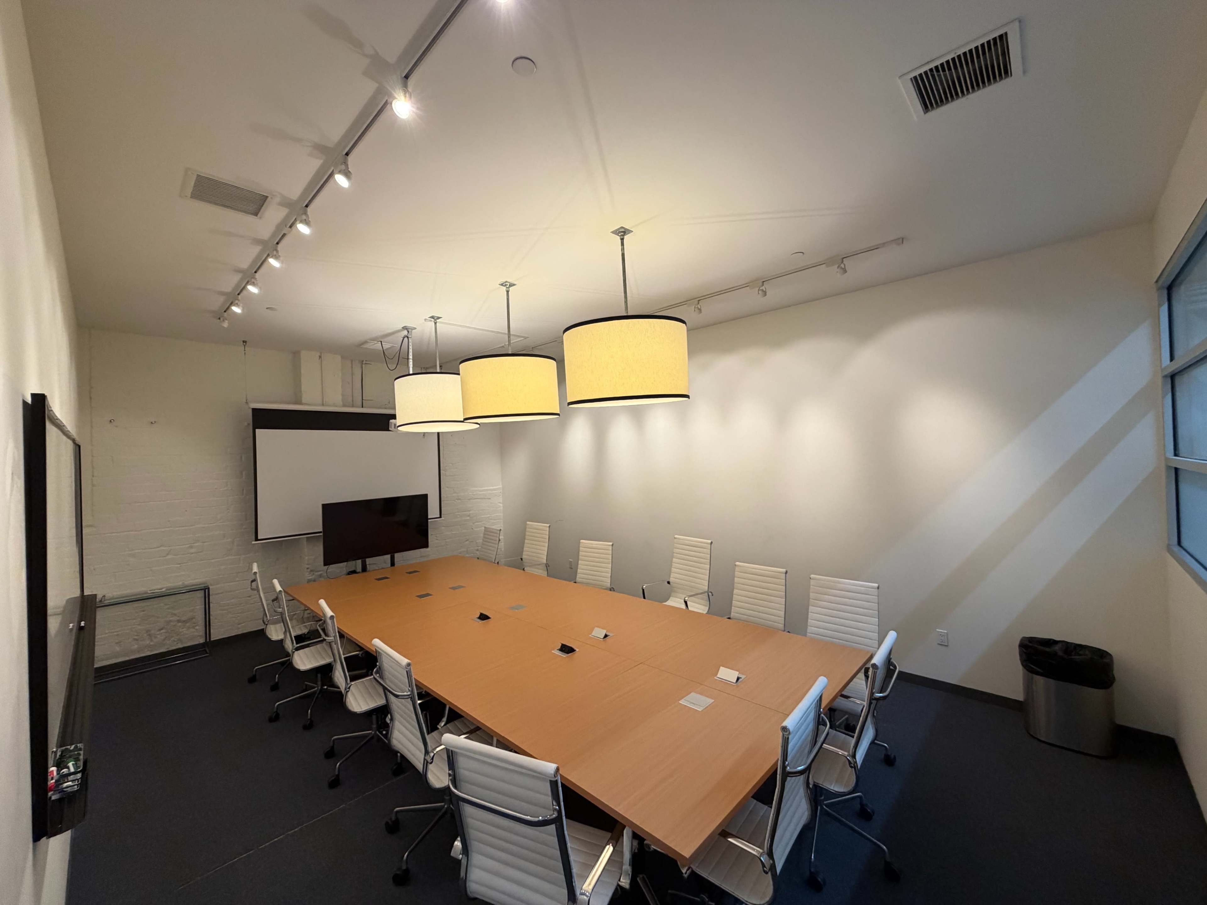 The image shows a modern conference room with a long wooden table surrounded by white chairs, featuring overhead lighting and a large screen at one end.