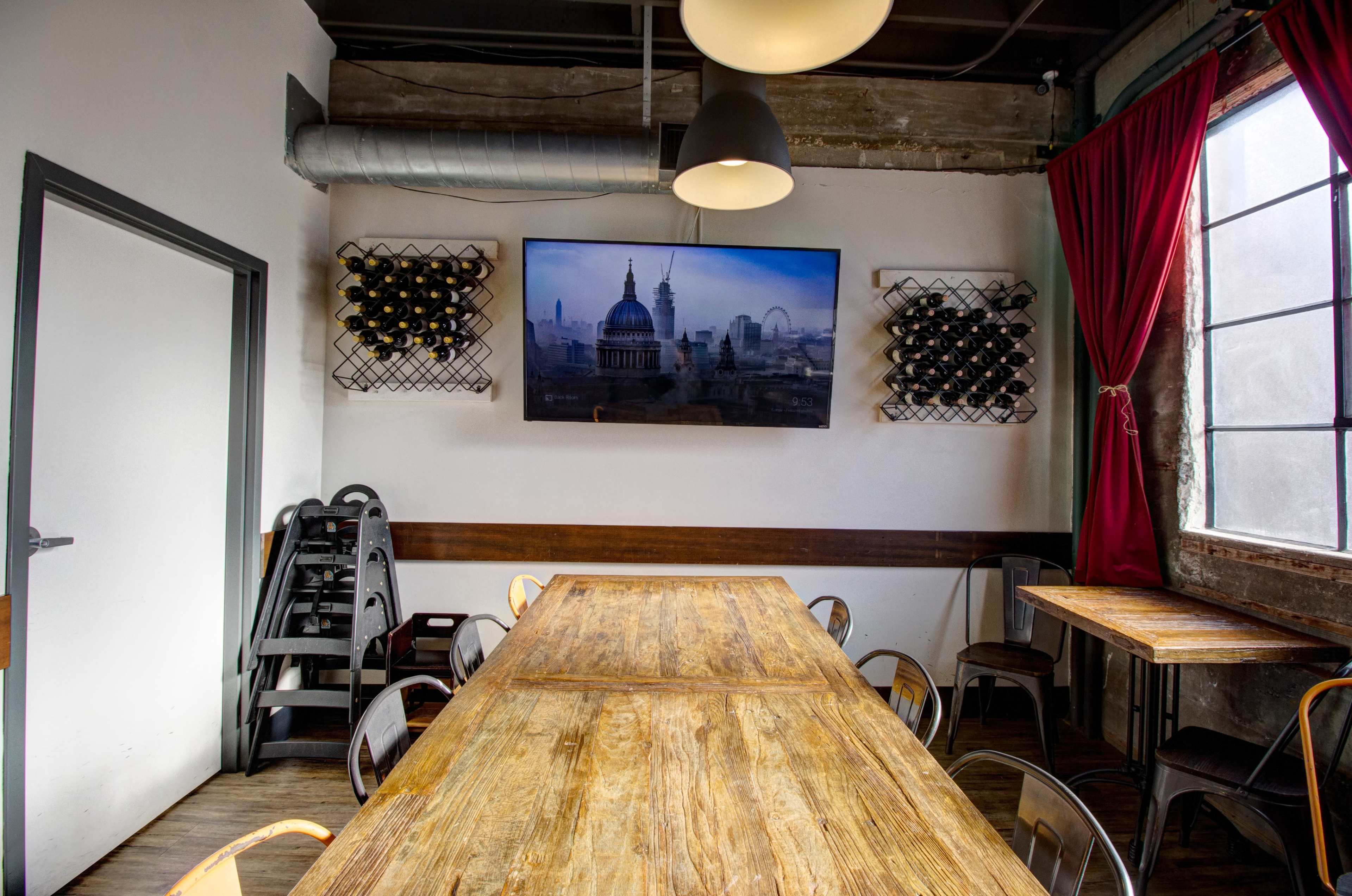 A spacious dining area features a long wooden table, metal chairs, and a large wall-mounted screen displaying a cityscape, with wine racks on either side.