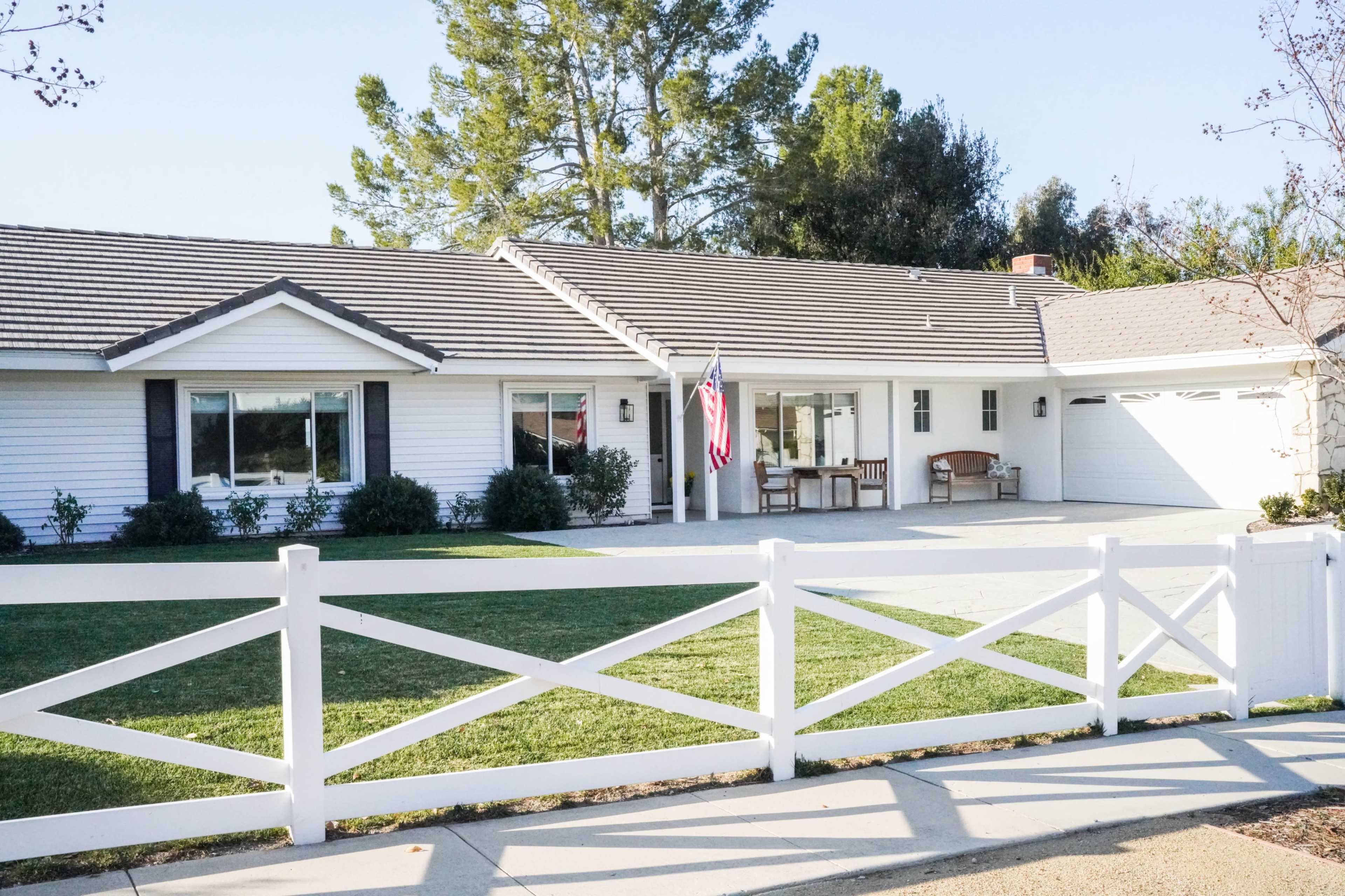 A single-story house with white siding and a grey roof features a flagpole with an American flag in the front yard, surrounded by a white fence.