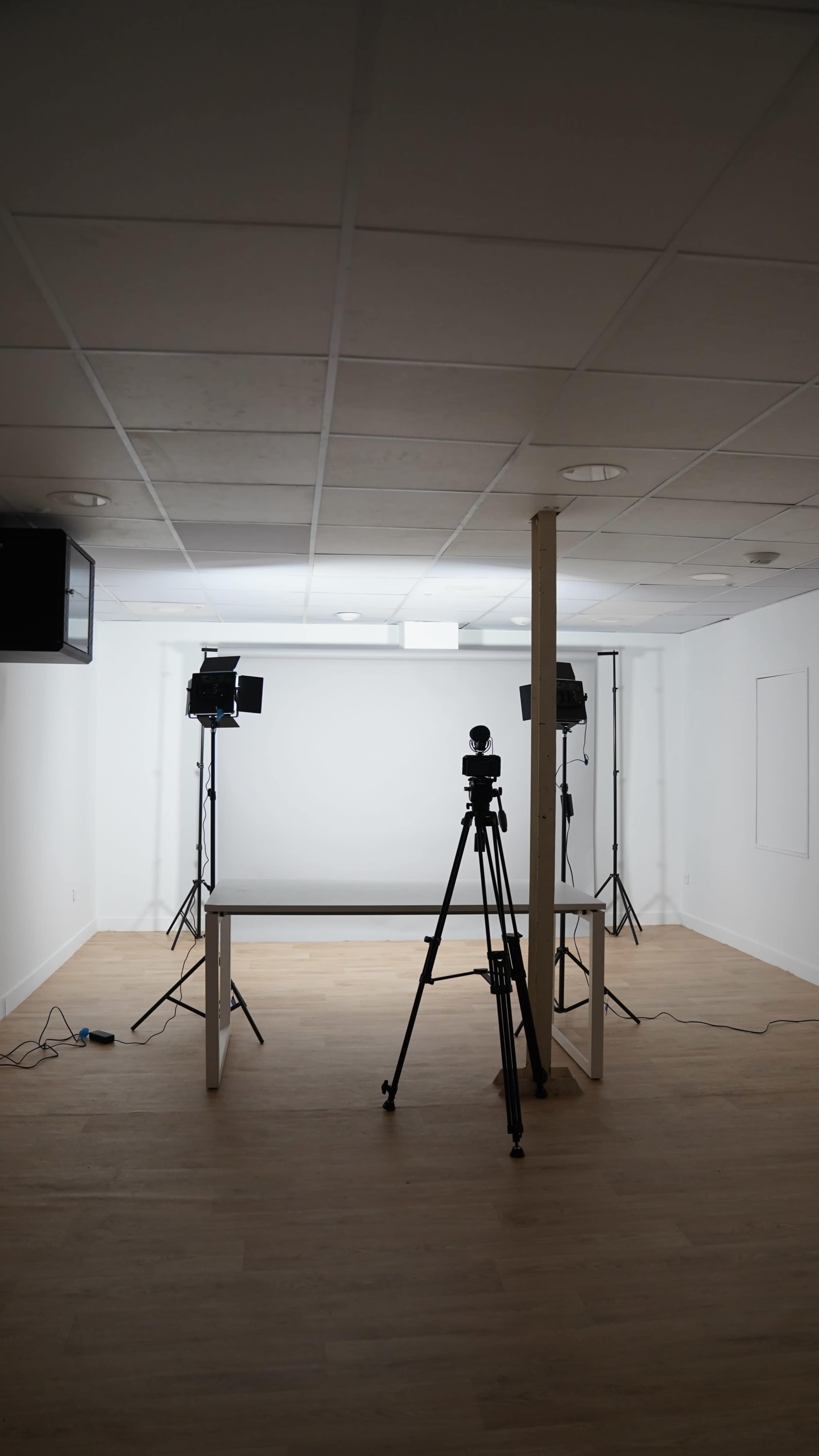 The image shows a sparsely furnished photo studio with two tripods holding cameras and studio lights positioned around a central table.